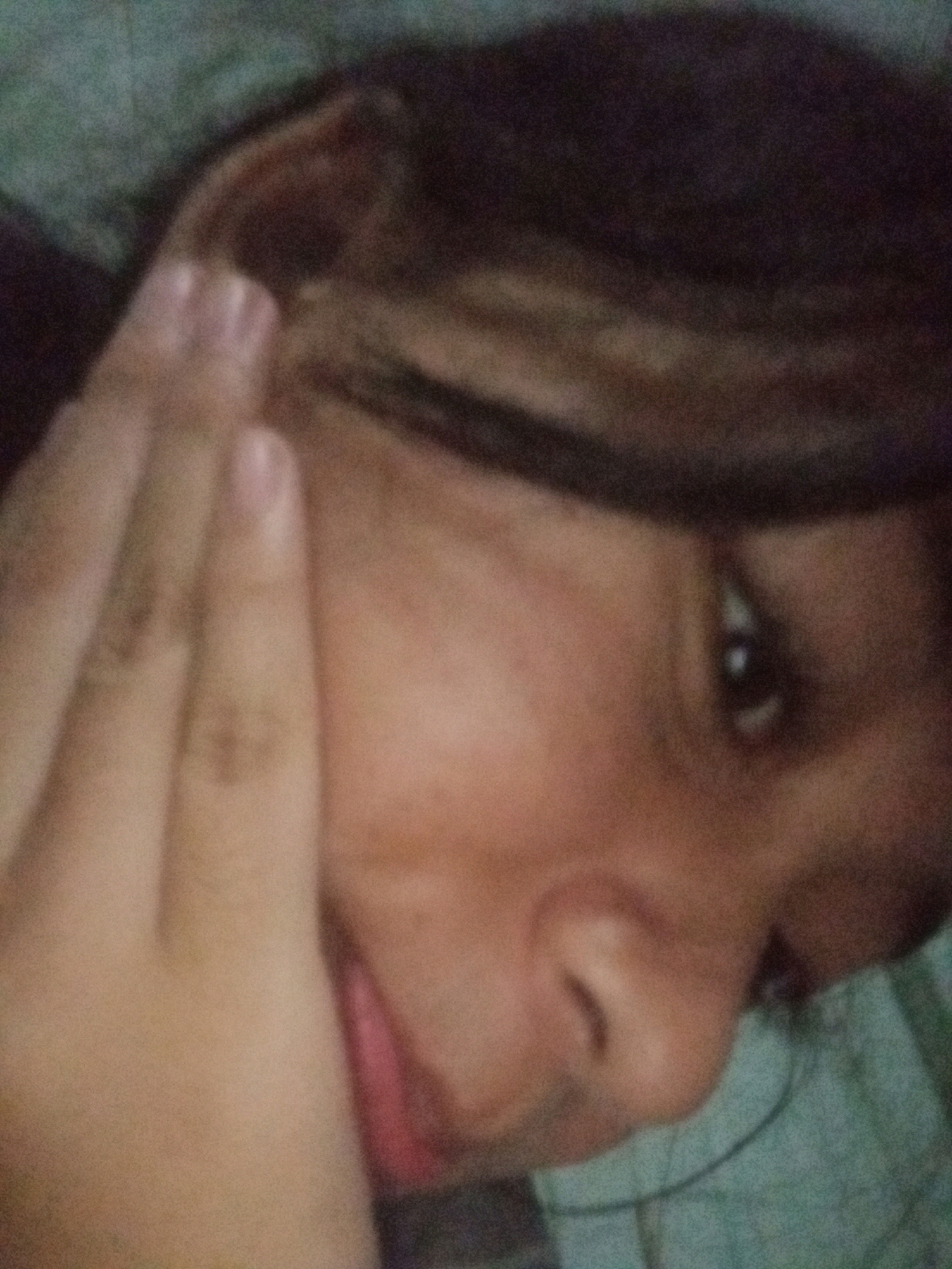 The image is a close-up, low-resolution photograph of a person's face, with their hand resting on their cheek. The person appears to be of Indonesian ethnicity. The background is out of focus and shows a textured surface, possibly a blanket or clothing, in shades of green and brown. The lighting is dim, and the image has a grainy quality. There is no discernible information about the location in Badas, Indonesia.