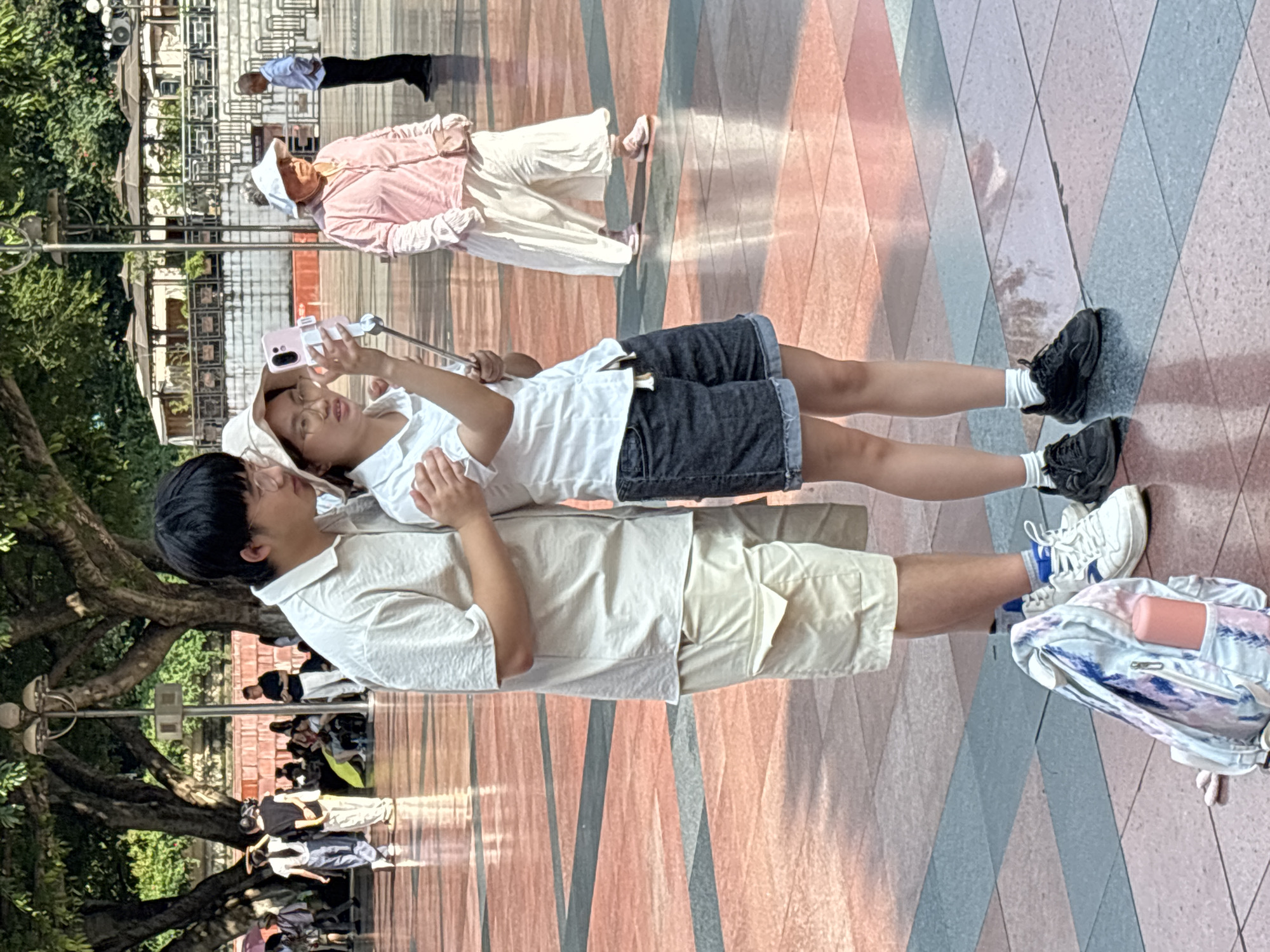 In Yuzhong District, China, two young people, a male and a female, are standing on a paved plaza. The male, wearing glasses, a light-colored short-sleeved shirt, and tan shorts, has his arm around the female. The female, wearing a white short-sleeved shirt, denim shorts, and a straw hat, is holding a selfie stick with a pink smartphone and looking at the screen. In the background, other people are walking and sitting. To the left of the young couple, a backpack with a pink water bottle inside rests on the ground. The plaza has a red and gray checkered pattern. The area is shaded by large trees.