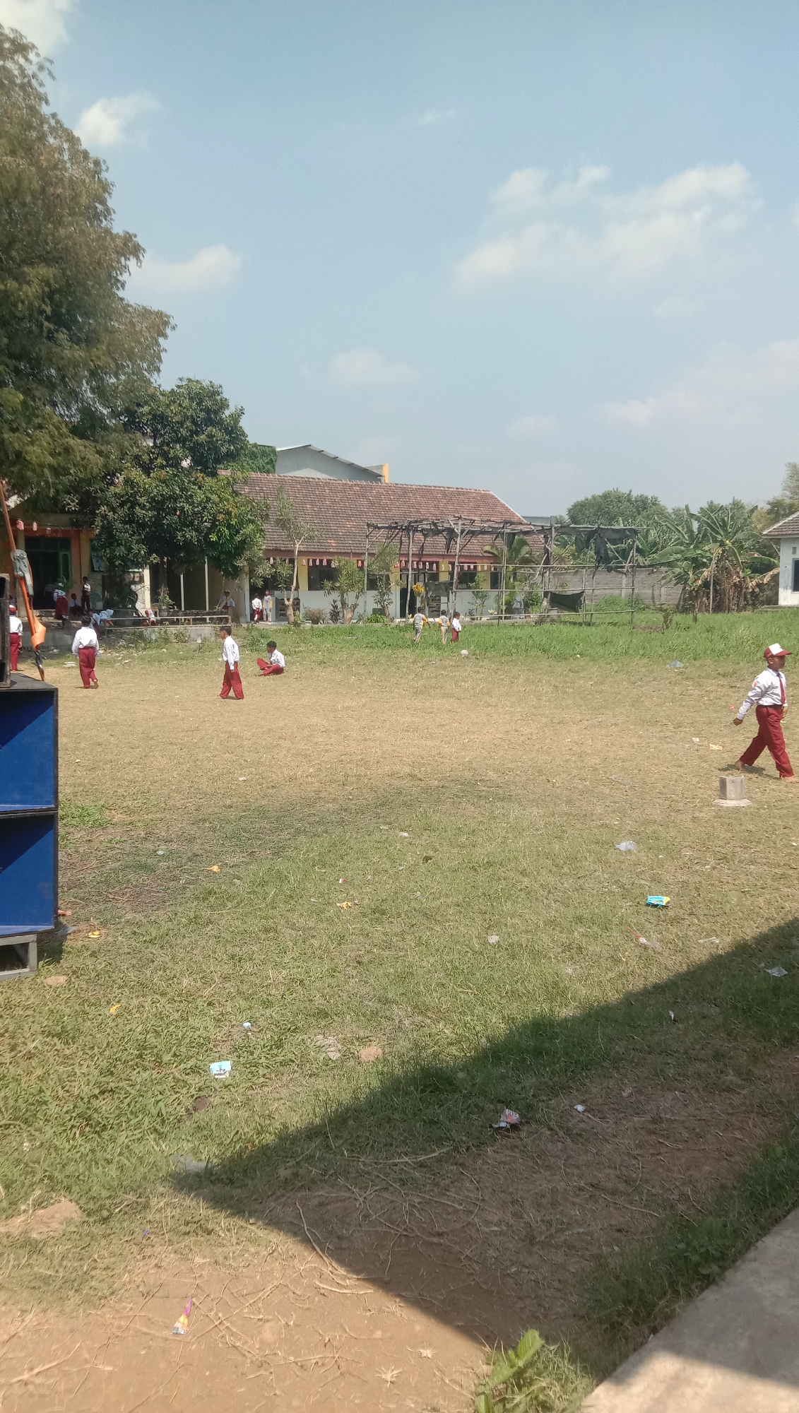 This is an outdoor scene of a schoolyard in Karang Anyar, Indonesia, under a clear blue sky with scattered white clouds. The ground is covered with dry, patchy grass. In the foreground on the left is a large, blue speaker. Further back, a group of children, presumably students, are scattered across the field, some standing and others sitting. They are wearing white shirts and red pants or skirts, typical school uniforms. Behind the field is a building with a red-tiled roof, a covered walkway supported by wooden posts, and several trees. The overall impression is of a sunny day at a school.