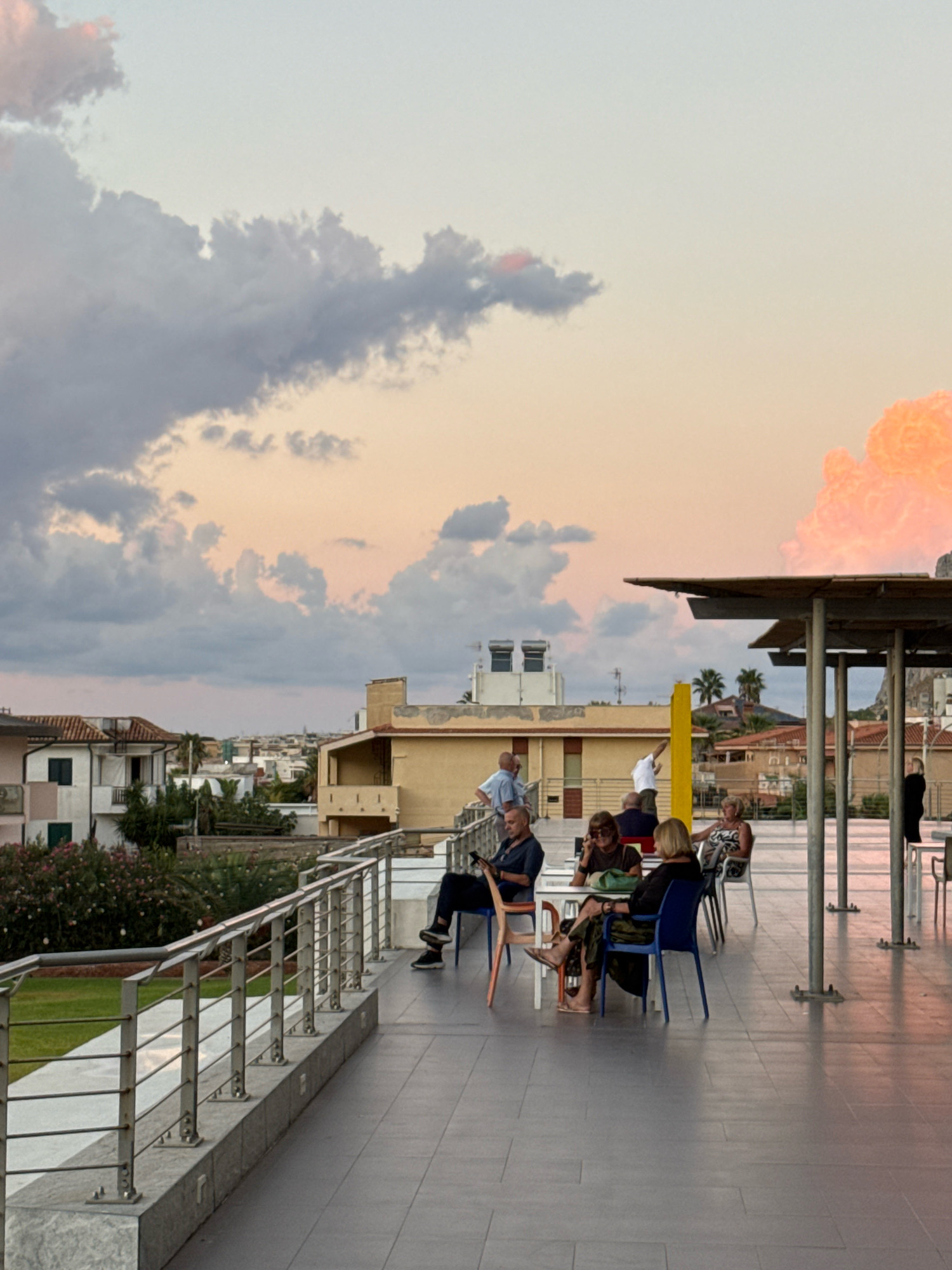 A rooftop terrace in Capaci, Italy, overlooks a town with buildings and a cloudy sky at sunset. Several people are seated at tables, some engaged in conversation, others looking at their phones. A few individuals stand by a railing, taking in the view. The scene suggests a relaxed evening atmosphere, possibly at a restaurant or cafe.