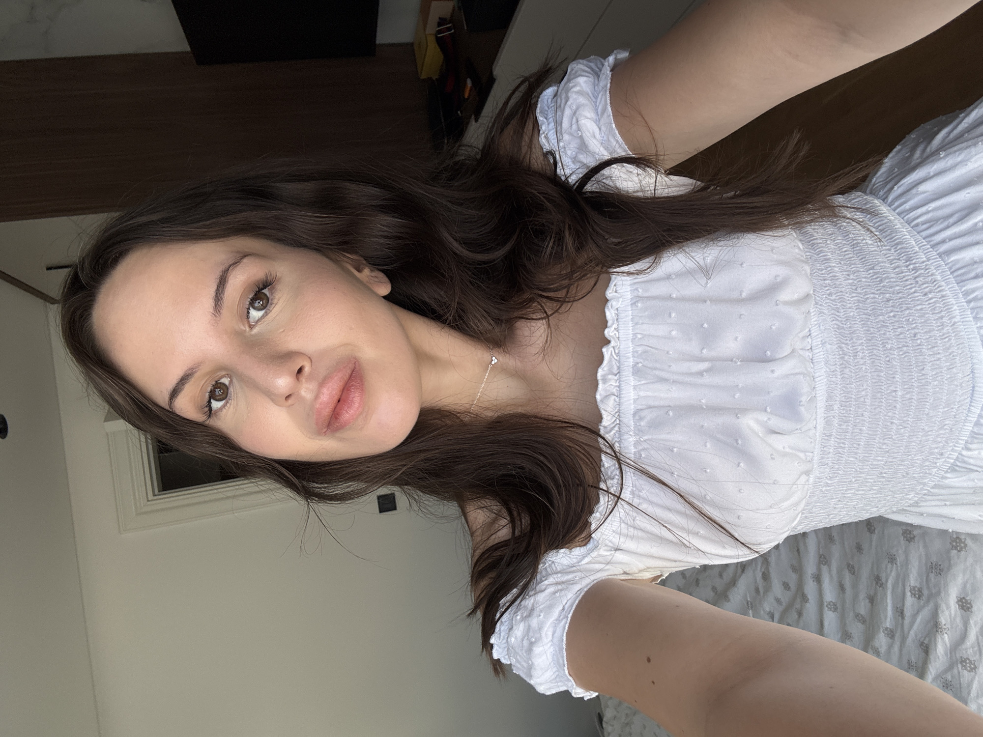 A young woman with long, dark, wavy hair is taking a selfie. She is wearing a white off-the-shoulder dress with a textured pattern and smocked bodice. A delicate silver necklace with a heart pendant is visible around her neck. Her makeup is subtle, with emphasis on her eyes and lips. The background appears to be a bedroom, with a bed visible in the lower left corner and a wooden closet or wardrobe to the right. Sunlight streams in from an unseen window, illuminating her face and right arm. The overall impression is a candid, relaxed moment captured indoors.