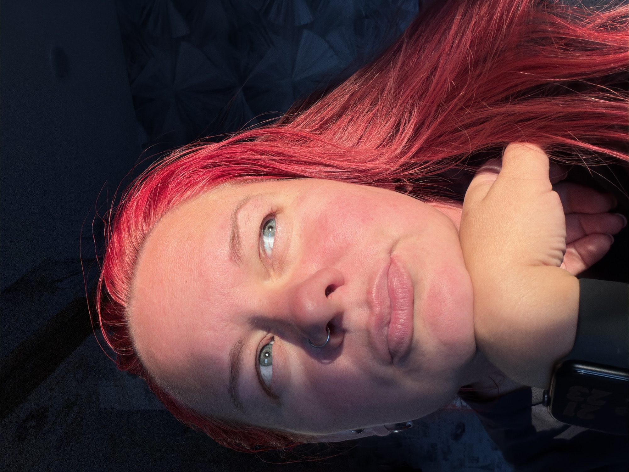 A close-up, head-and-shoulders portrait shows a person with bright red hair, resting their chin on their hand. The subject is looking off to the right, with their right eye slightly more visible. Sunlight illuminates the left side of their face, highlighting rosy cheeks and a nose ring. The background is dark and textured, with some diagonal shadows visible at the top left. A smartwatch is visible on the subject's left wrist. The overall impression is one of a casual, indoor setting, possibly a home. There is no indication of the city of Chaikovsky, Russia in the image.