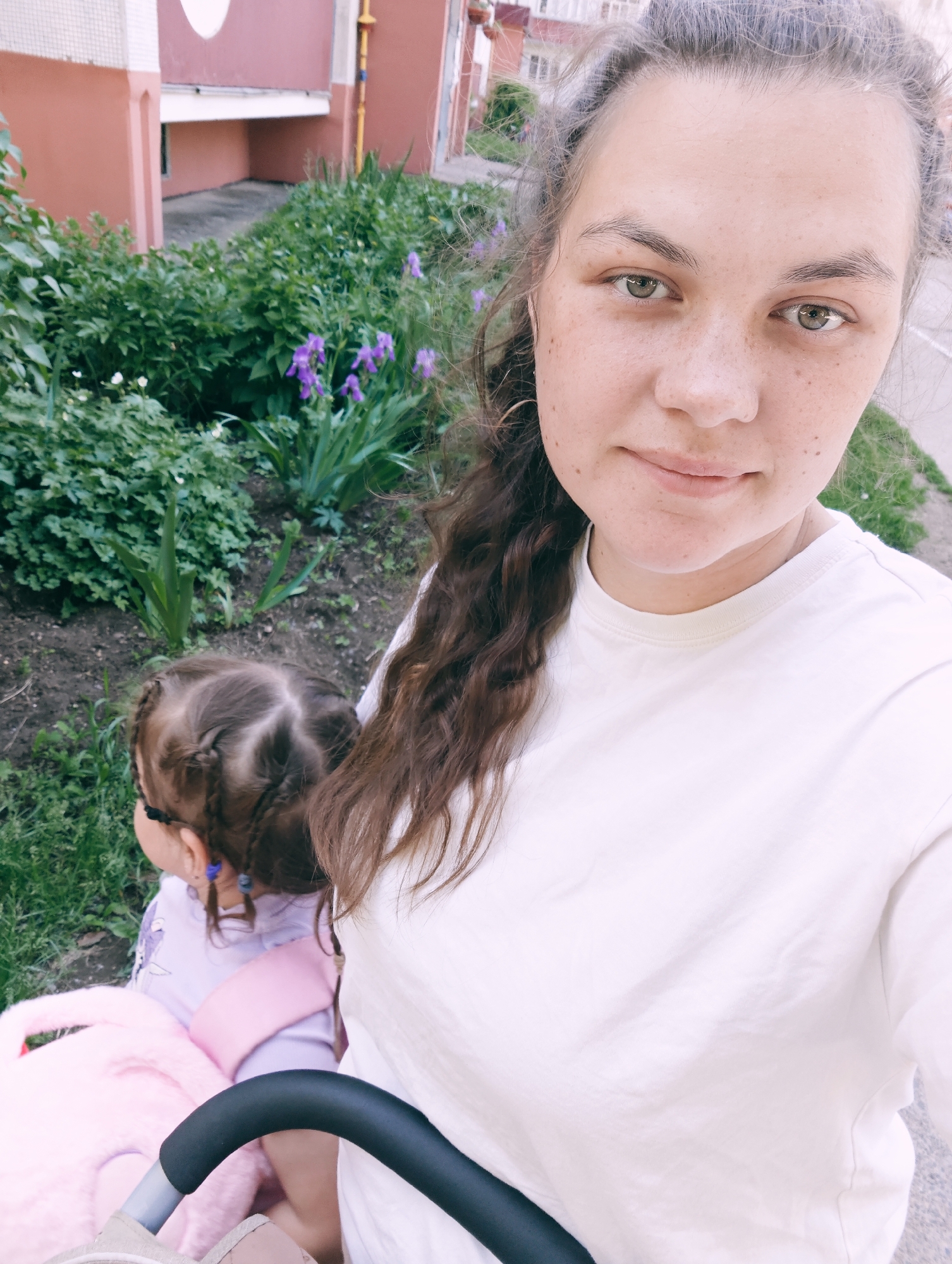 A woman and a young child are outdoors near a building. The woman is in the foreground, facing the camera with a slight smile. She has brown hair pulled back and is wearing a white t-shirt. She has freckles on her face and neck. Partially visible behind her and to the left is the child, facing away from the camera. The child has brown hair styled in braids and is wearing a purple shirt and a pink harness. A pink blanket and stroller handle are visible in the lower part of the frame. The background shows a building with pink and beige walls, a yellow pipe, and green bushes with purple flowers. The overall setting appears to be a residential area in the city of Chaikovsky, Russia, suggesting a casual outing in a park or yard.