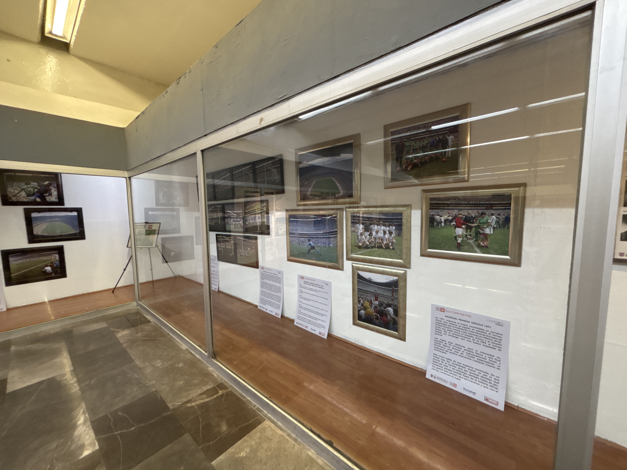 The image depicts an interior space, possibly a museum or exhibition area, in Mexico City, Mexico. The foreground features a polished, tiled floor with dark gray and brown patterns. A glass enclosure separates the foreground from a wooden floor that is an exhibition area. On the back wall of the glass enclosure, several framed photographs and informational posters are mounted. The pictures primarily seem to be sports-related, specifically soccer. The text on one poster is partly visible and seems to discuss "MUNDIAL FEMENIL MÉXICO 1971," referring to a women's soccer world tournament in Mexico in 1971. There is an artificial light fixture at the top left of the image. The walls are painted in neutral colors, likely white or light gray.