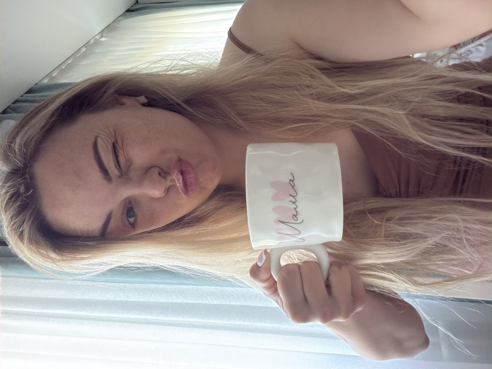 A woman is holding a white mug that has the Russian word "Mama" written on it in cursive. She is making a kissy face, winking one eye. The background is a window. The photo's location is unknown.