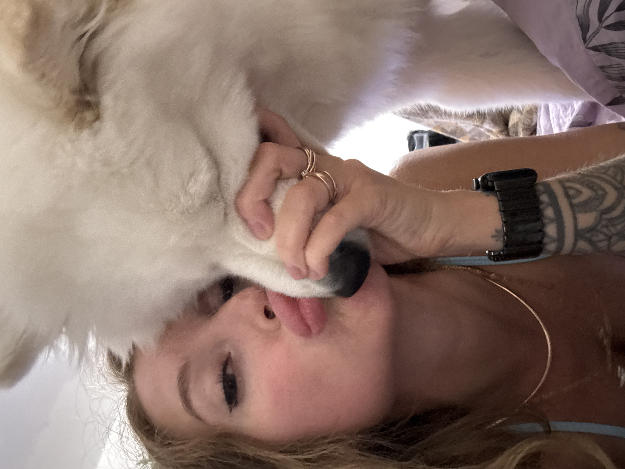 A woman is lying down, holding a large, white dog's face near her own. She is sticking out her tongue. The dog's nose is black. The woman is wearing rings on her hand, a black watch on her wrist, and a gold necklace. Her arm has a tattoo. A patterned blanket is visible in the background. It is not possible to verify the location in the city of Chaikovsky, Russia, from the given image.
