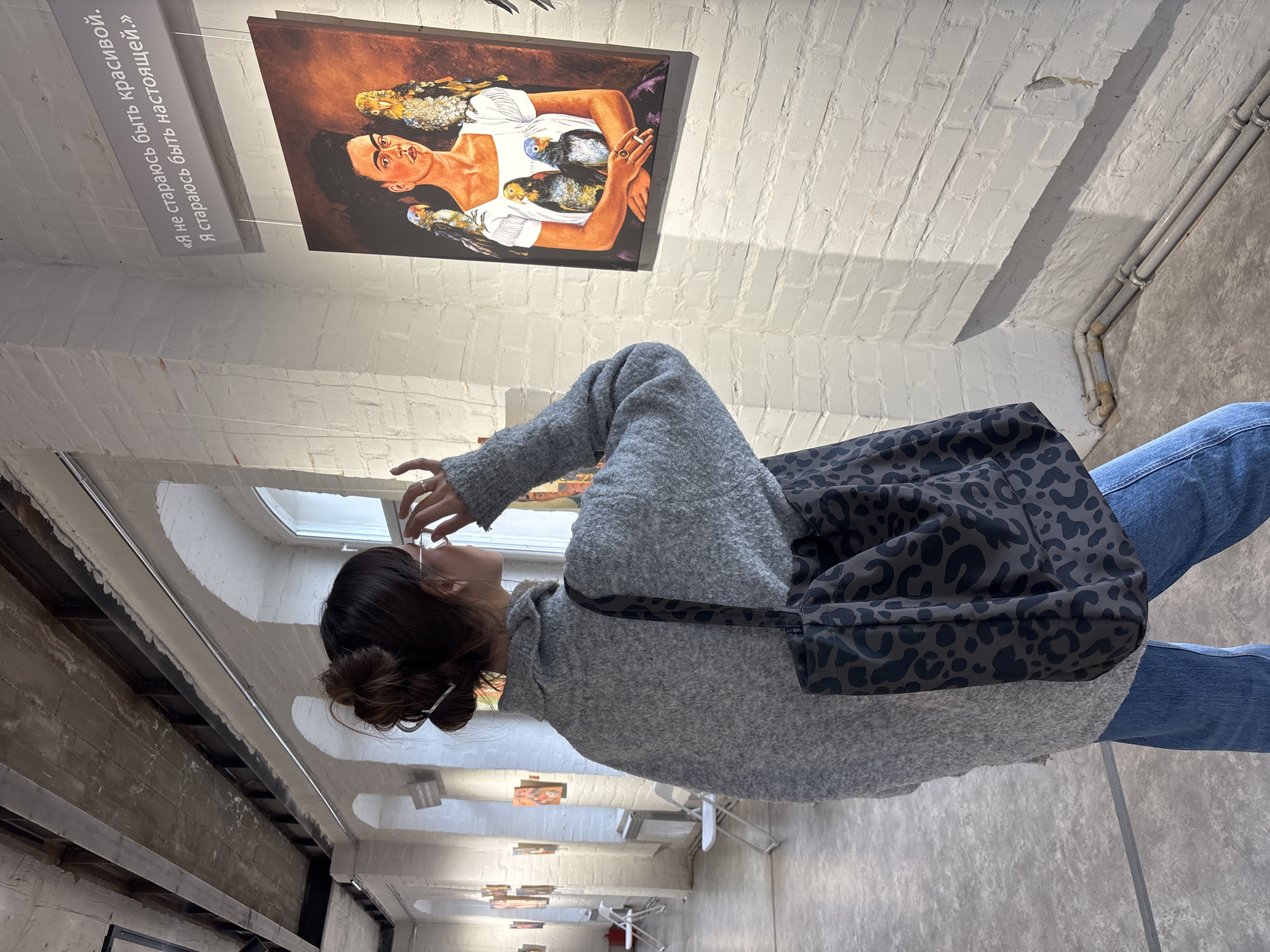 A young woman with her hair in a bun is standing in an art gallery, looking up at a painting. She is wearing blue jeans, a grey sweater, and a black and grey animal print bag. The painting she is looking at depicts a woman with birds. The gallery has white brick walls, white ceiling, and concrete floors. A sign with Cyrillic text is next to the painting. Other artworks are visible along the corridor. This image appears to be taken in Krasnoyarsk, Russia, based on the text on the sign.
