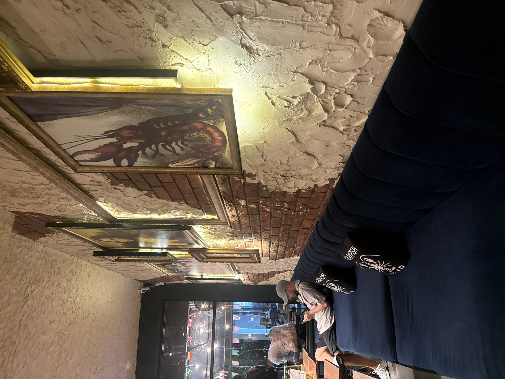 The image depicts the interior of a restaurant or bar, characterized by a unique blend of rustic and modern elements, set in Kuta, Indonesia. The walls feature a textured, off-white plaster finish, with sections of exposed red brick, particularly higher up, creating a distressed aesthetic. A long, dark blue velvet-upholstered bench or booth runs along one side, furnished with two black square pillows.
Seated on the bench is a man, engrossed in his phone. He wears a light-colored t-shirt, dark shorts, and a baseball cap. His reflection is visible in the large window or glass door to his left, which looks out onto a street or outdoor area, reflecting interior lights and indistinct shapes. The overall lighting is dim and warm, suggesting an evening setting.
The wall above the bench is adorned with several framed pictures, some illuminated by concealed strip lighting. A prominent framed artwork depicts a large red lobster. Another frame, partially visible, appears to hold a classic portrait, possibly the Mona Lisa. The black pillows on the bench clearly display the word "CRAYFISH" (and "CRAWFISH" on another) alongside an illustration of a crawfish or lobster, indicating a possible theme for the establishment. Two security cameras are mounted near the top left corner of the window frame. The scene conveys a relaxed and intimate atmosphere.