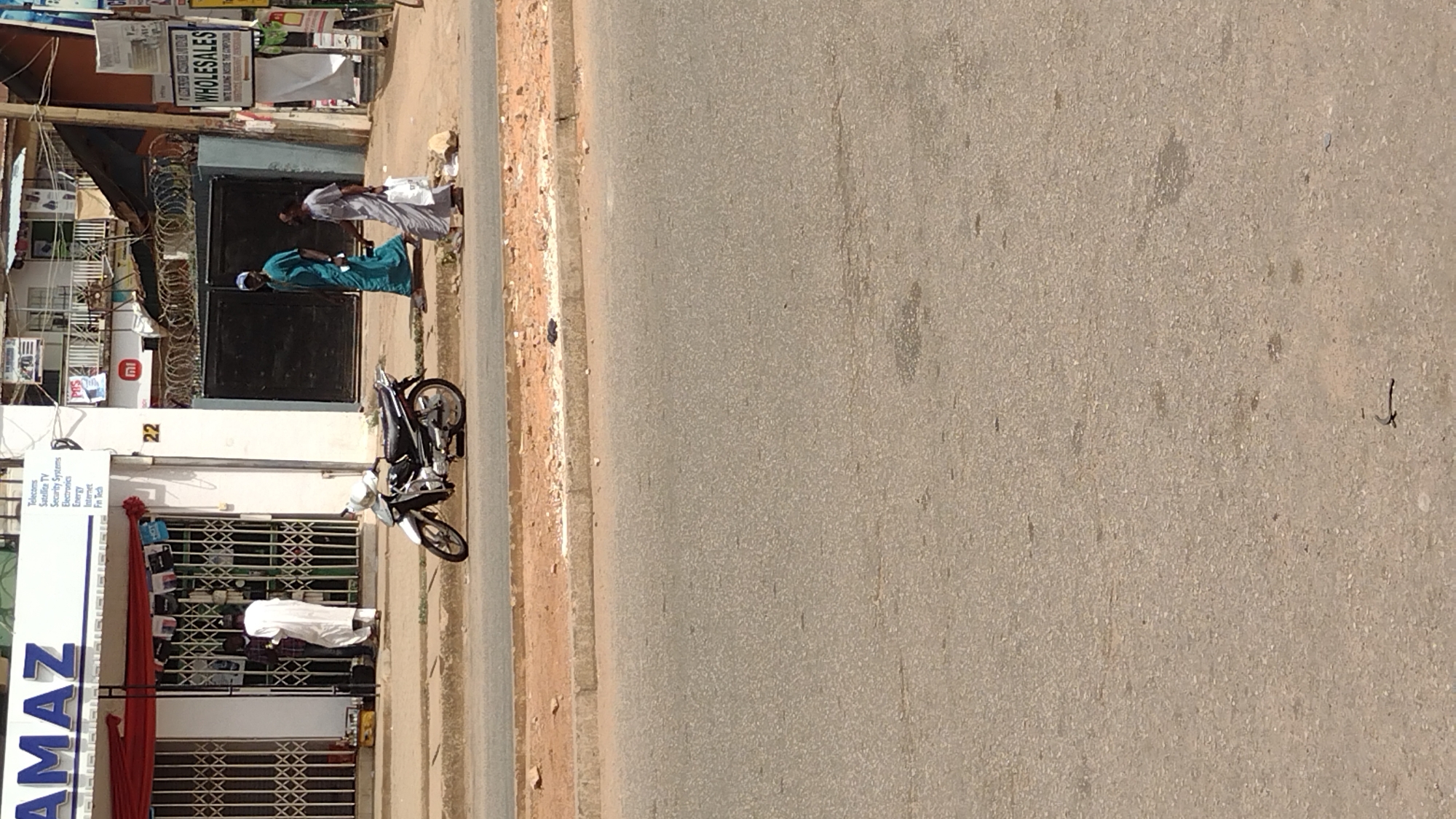 Here is a description of the image:
The image shows a street scene, likely taken in Jos, Nigeria, indicated by the business signs. A wide asphalt road occupies much of the frame. The roadside includes a sidewalk and storefronts.
A business with a sign reading "AMAZ" offers services including Telecoms, Satellite TV, Security Systems, Electronics, Energy, Internet, and Fin Tech. A number "22" is also visible on the building. Another sign displays "WHOLESALES, WHITE BALING INSIDE THE COMPOUND".
Several people are visible. One man wearing a white robe stands in front of the "AMAZ" storefront. Two other men in traditional garments are walking away from a gate.
A parked motorcycle stands near the businesses. Security features such as razor wire are above the gate.