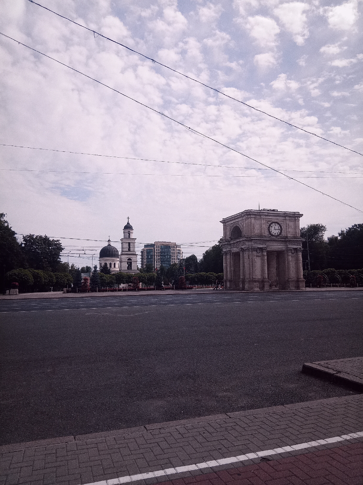 The image captures a wide view of a city square in Chișinău, Moldova. In the foreground, a paved road with white lane markings is visible, leading into the distance. To the right, a sidewalk with brick paving and a white line runs along the edge of the road. In the mid-ground, a large, stone triumphal arch, the Arcul de Triumf, stands prominently. It features classical architectural elements, including columns and an arched passageway, and a clock face is affixed to its facade. To the left of the arch, a white church with a dark dome and a bell tower topped with a cross is situated. Behind the church, a modern, glass-fronted building rises. The scene is framed by a line of trees and bushes on either side of the square. Overhead, multiple power lines crisscross against a cloudy sky. The overall impression is that of a public landmark area in a city.