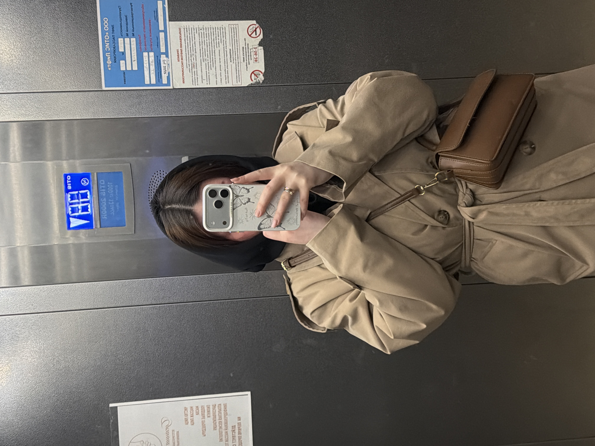 An individual is captured in a mirror selfie within an elevator, their image appearing rotated in the photograph. The person, mostly visible from the chest upwards, wears a light beige or tan trench coat and a dark head covering or scarf. They are holding a smartphone with a white, butterfly-patterned case, obscuring part of their face while taking the picture. A ring is visible on their right hand. A small, dark brown leather crossbody bag with a gold-colored clasp rests on their coat.
The elevator interior features brushed metal walls. A blue illuminated digital display on the wall shows what appears to be the letter "B" (possibly indicating a basement or ground floor) with a downward-pointing arrow, and some Cyrillic text including "BITO" and "1000 BITO". Several notices and informational signs are affixed to the elevator walls. Some of these papers appear worn or slightly torn. Visible Cyrillic text includes what seems to be a company name "«ТФИП ЭНТО» ООО", the number "3-20-38", and various other words and phrases in Russian, some of which are difficult to decipher clearly due to their condition or reflection. The overall setting indicates an indoor public or commercial building in Chaikovsky, Russia, given the Cyrillic script.