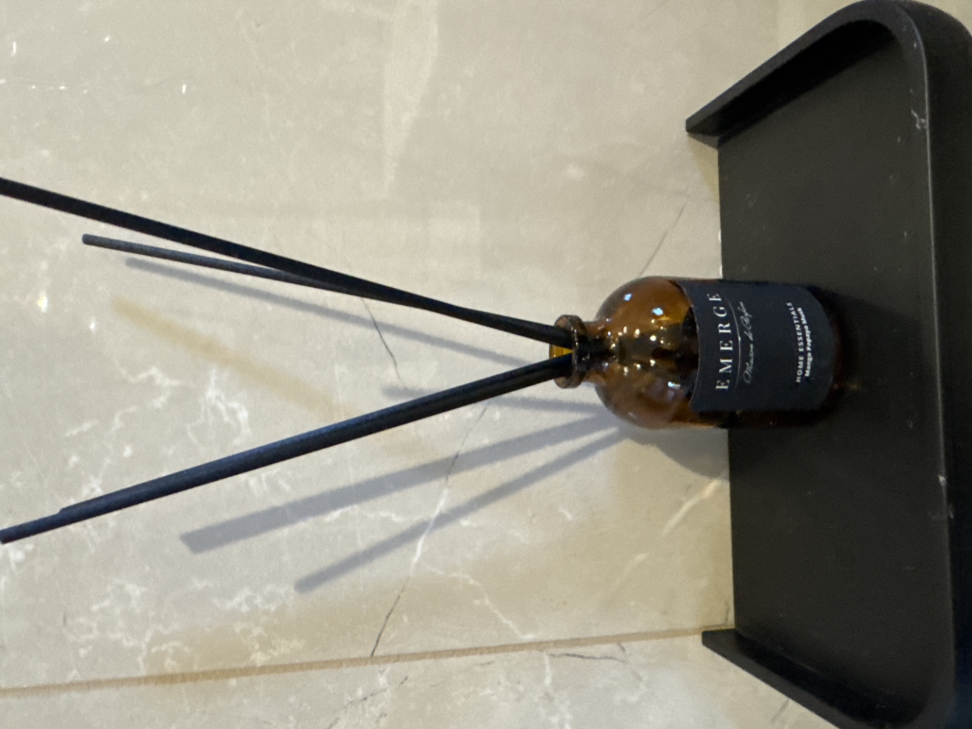 A close-up, eye-level shot shows a reed diffuser with a dark amber glass bottle and three dark, slender diffuser sticks protruding from the top. The bottle has a black label with white text that reads "EMERGE Maison de Parfum" and "HOME ESSENTIALS Mango Papaya Musk". The diffuser sits on a black, rectangular tray with a slightly curved edge. The tray is positioned in front of a wall with a light-colored, marbled texture. The lighting casts shadows of the diffuser sticks onto the wall. The image does not contain any information that would indicate its location in Soloneshnoye, Russia.
