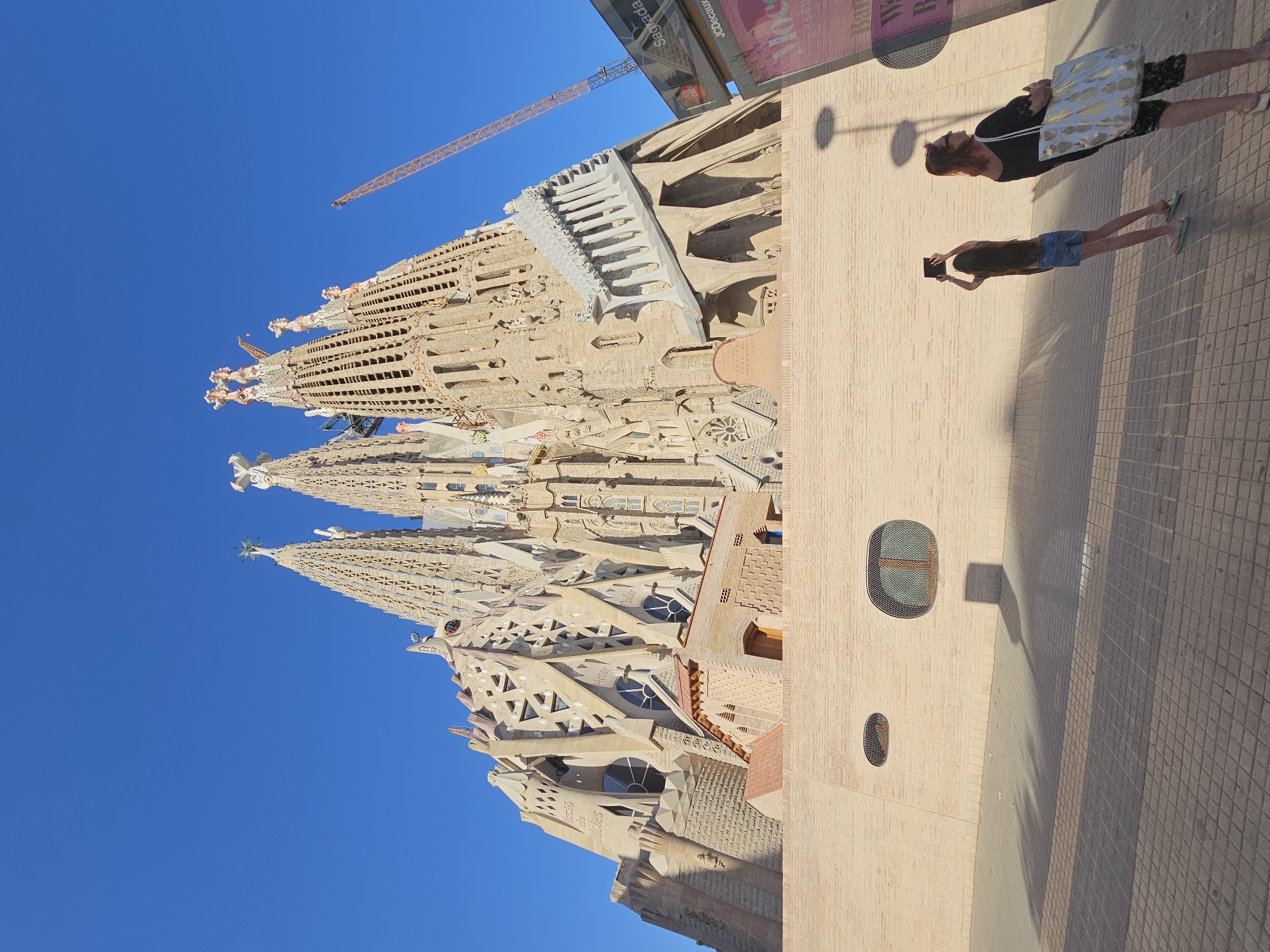 This media file captures a perspective view of the Sagrada Familia basilica in Barcelona, Spain, against a clear blue sky. The intricate and unfinished spires of the basilica dominate the upper left portion of the frame, showcasing its unique architectural style. A construction crane is visible in the background, indicating ongoing work on the basilica.
In the foreground, on the right side of the image, a light-colored brick building with small, circular windows stands adjacent to the basilica. Two individuals are present on the tiled ground in front of this building. One person, wearing a black top and patterned shorts, is looking upwards and to the left, holding a light-colored bag. Another person, wearing a blue top and shorts, is holding a device, possibly a phone or tablet, and appears to be taking a picture or recording. Their shadows are cast on the ground, suggesting it is daytime, likely late afternoon given the angle of the shadows. The overall atmosphere is one of tourism and admiration for the iconic landmark. A partial view of a banner with text is visible at the top of the brick building, but the text is largely illegible.