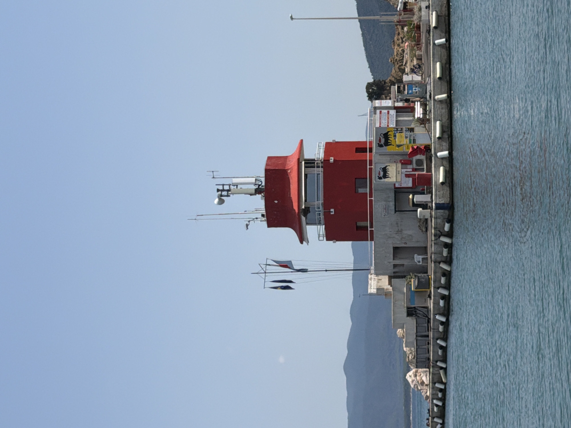 A red, multi-story building with a distinctive red, pagoda-style roof sits on a concrete pier extending into blue water. The building has several windows and is topped with communication antennas. Flags fly from a nearby flagpole, which has three flags; two dark blue and one with red, white, and blue vertical stripes. The pier is lined with white fenders. Behind the building, a rugged, rocky shoreline with green vegetation can be seen, backed by hazy mountains under a clear blue sky. The pier area also contains a gas station with pumps and signage, a white plastic chair, and a bicycle. The building appears to be a lighthouse or a maritime control tower.