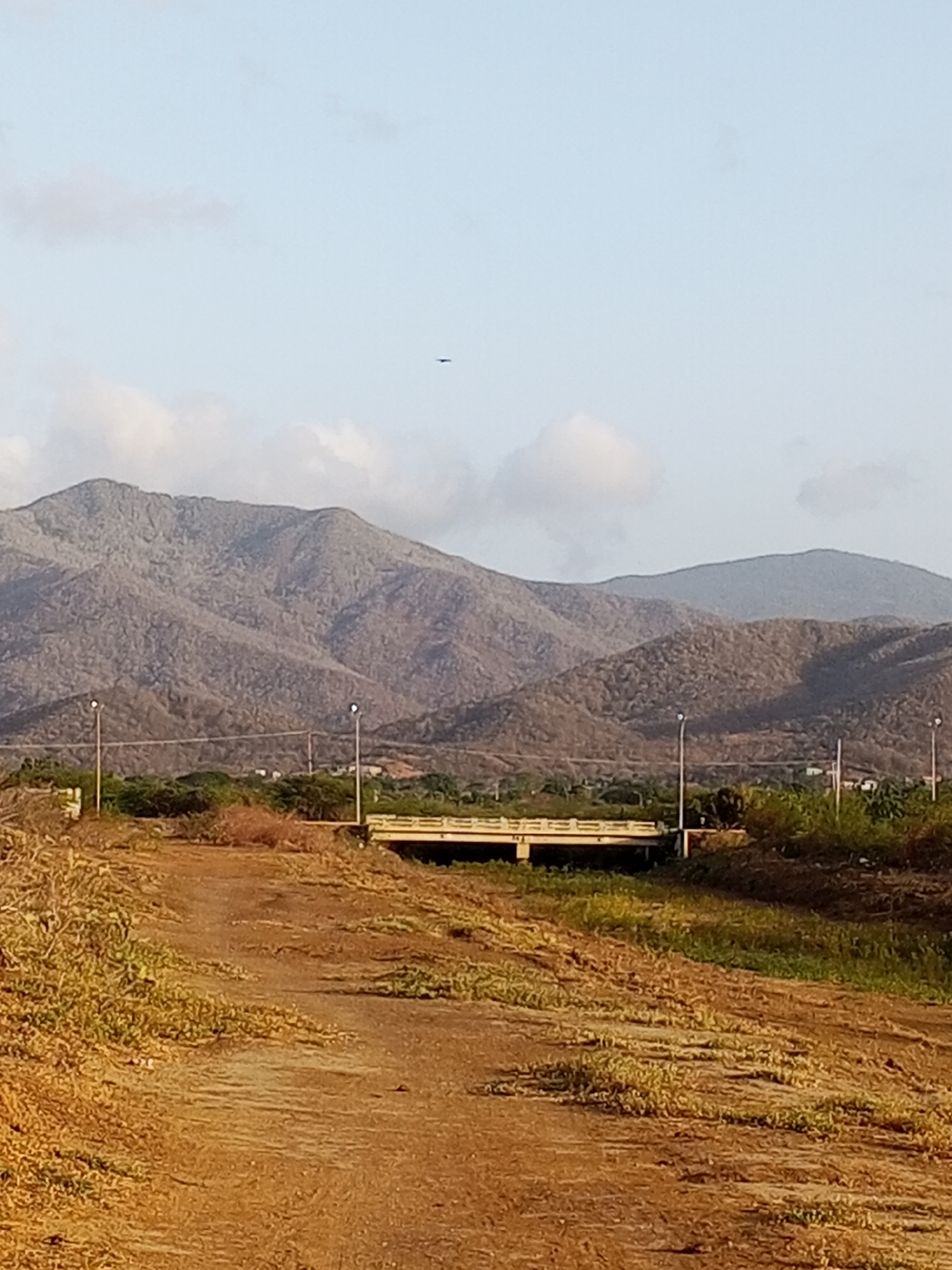 The image shows a landscape with a dirt road in the foreground leading toward a bridge. The road is covered with sparse vegetation. In the midground, there is a concrete bridge with several light poles along its side. Behind the bridge, there are trees and possibly some buildings. In the background, a range of hills or mountains dominates the view, under a sky with some clouds. The location appears to be outside the main urban area, possibly on the outskirts of a city or town.