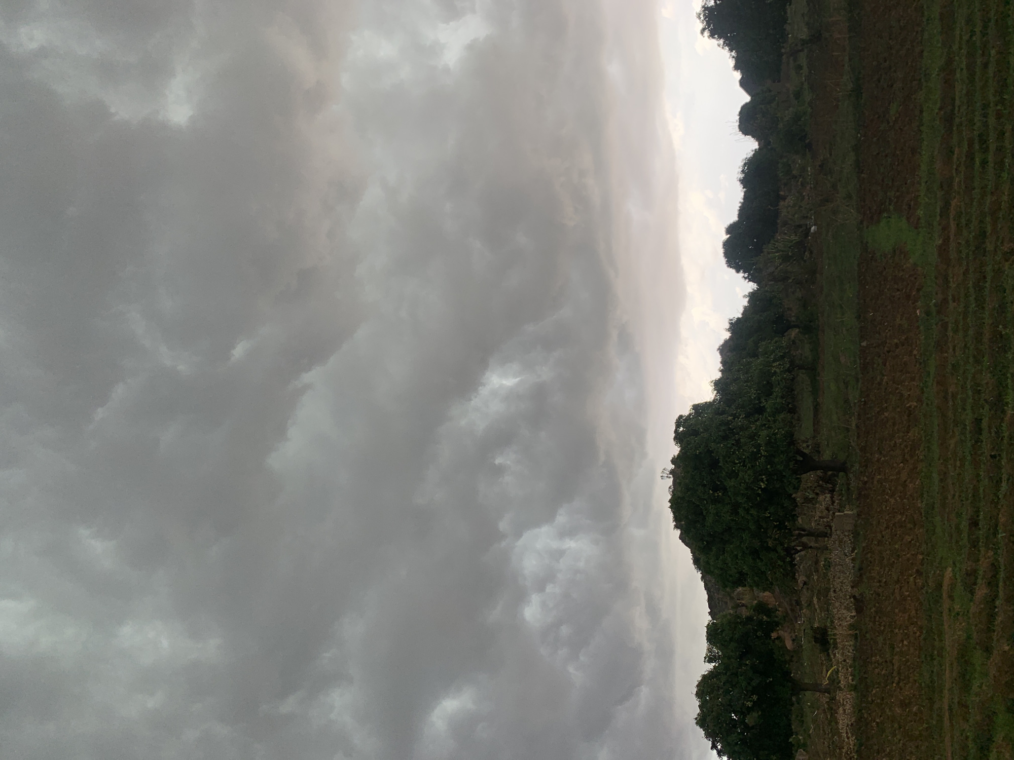The image shows a landscape in Gwafan, Nigeria under an overcast sky filled with grey, heavy clouds. A strip of land runs along the right side of the image, featuring trees and vegetation. The ground appears to be a mix of cultivated fields and grassy areas. There are no visible people or animals. The overall environment is somber due to the stormy-looking sky.