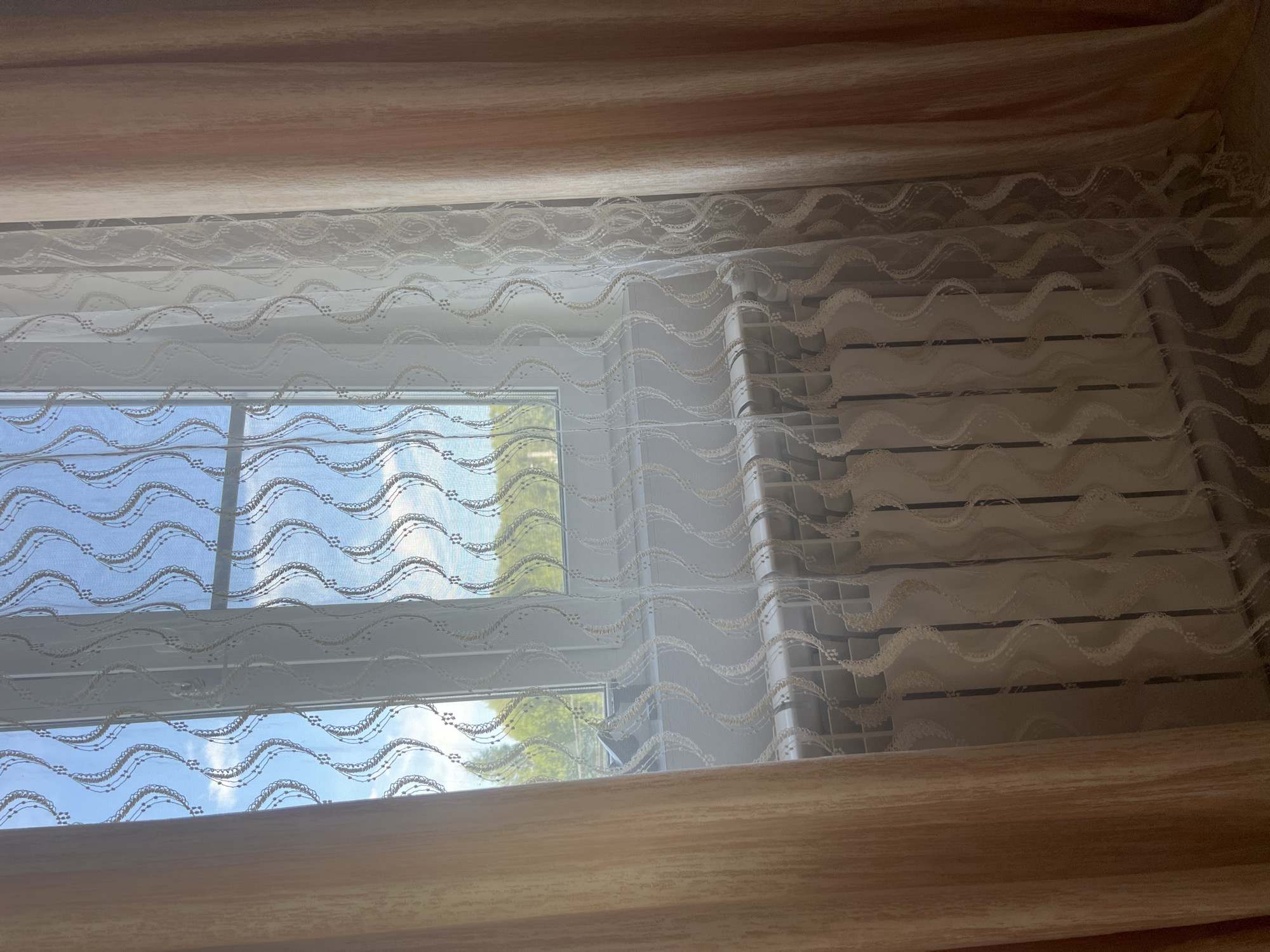 The image shows a window with curtains and a radiator. The window is partly covered by a sheer white curtain with a wavy pattern, and to the left and right are thicker, peach-colored curtains. Through the sheer curtain, a view of trees and a blue sky with some clouds is visible. Below the window is a white radiator with a ribbed surface. The scene is indoors, likely in a room. There are no people or animals visible. The image does not provide any information or context to suggest a specific location in the city of Zhukov, Russia.