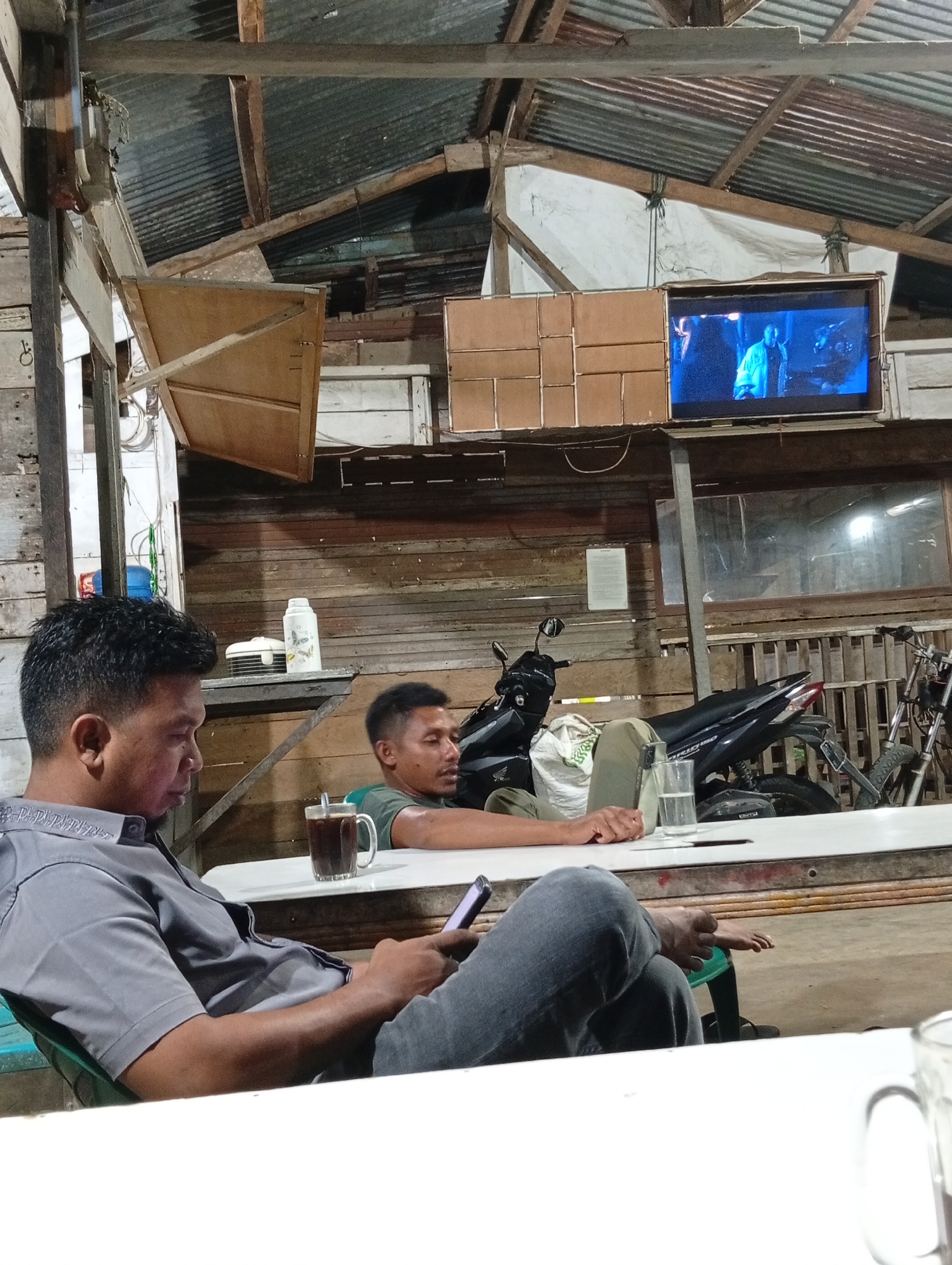 An interior scene in Lhokseumawe, Indonesia, depicts a rustic establishment, likely a cafe or communal space. The roof is constructed from corrugated metal sheets supported by wooden beams, with some sections covered by white tarpaulin. The walls are primarily made of horizontal weathered wooden planks.
Two men are seated at a long, white rectangular table. In the foreground on the left, a man with short black hair and a faded haircut wears a grey collared short-sleeved shirt with subtle embroidery and dark jeans. He is looking down at a black smartphone held in his right hand. A clear mug containing a dark liquid with a spoon rests on the table in front of him. His left forearm is on the table.
Behind him, to the right, a second man with short black hair and a dark green t-shirt is seated, leaning back slightly with his arms resting on the table. He looks towards the right. His bare feet are visible under the table near the foreground man's leg. A glass of water is on the table near his right hand.
High on the wall, to the right, a television screen is enclosed in a wooden frame, displaying a blue-toned scene with two figures. To the left of the television are several light brown rectangular panels arranged decoratively or as shelving. A white thermos and a small lidded container are on a shelf in the background.
Two motorcycles are parked in the background behind the table: a black scooter (possibly Honda) on the left and an older, partially visible motorcycle or bicycle on the right. Exposed wiring, a blue container, and various miscellaneous items are visible throughout the background, contributing to the informal and functional aesthetic of the space.