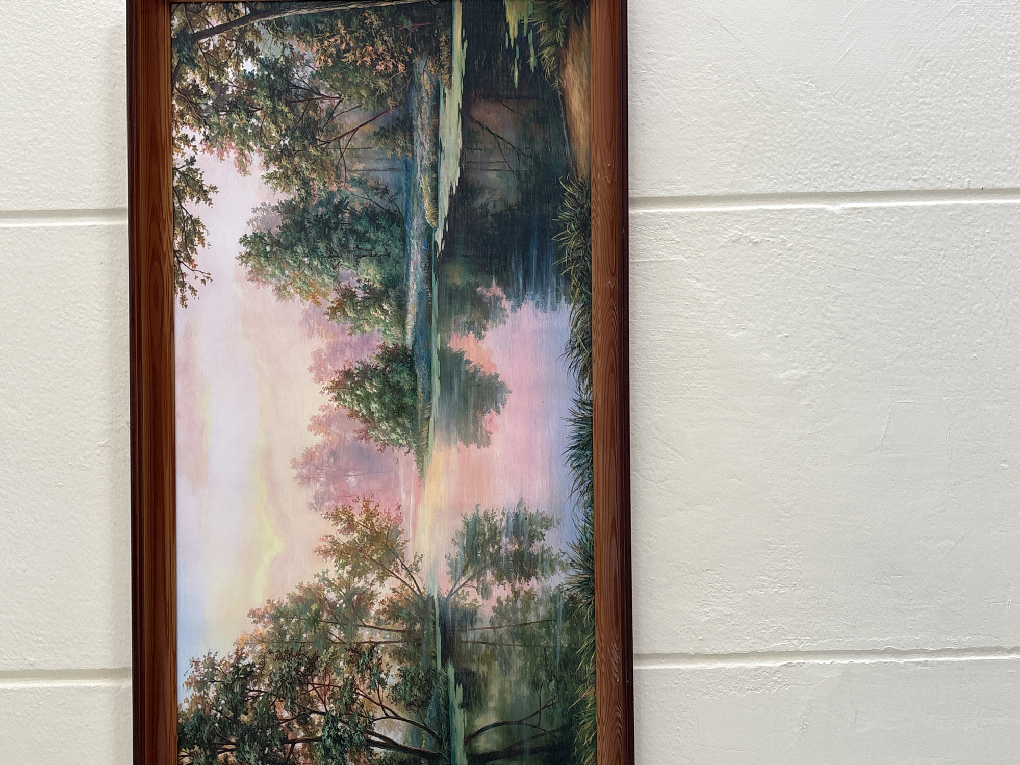 The image displays a framed painting depicting a serene landscape. The painting features a tranquil body of water, likely a lake or river, reflecting the soft hues of a sunset sky. Lush green trees with dark trunks line the water's edge, their branches creating a dense canopy. The sky is painted with soft pastels of pink, orange, and yellow, suggesting a peaceful twilight. The painting is framed in a dark wooden border. The artwork is hung on a textured white wall, divided by vertical lines, indicative of a building's interior. There is no discernible connection to the city of Vitsebsk, Belarus.