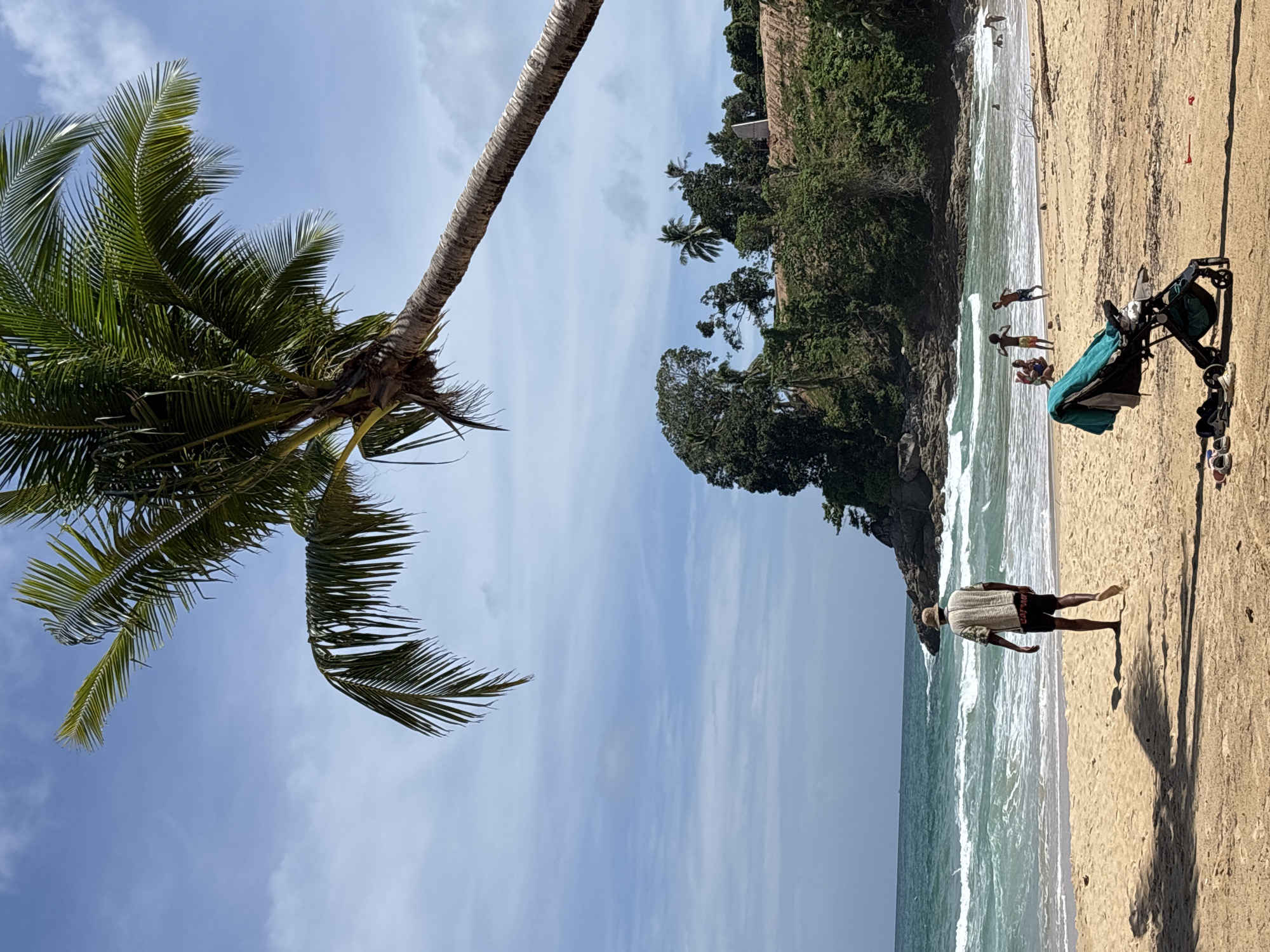 This outdoor shot captures a beach in Choeng Thale, Thailand. The image has been rotated, with the sky and a palm tree appearing on the left. The beach is sandy and meets the ocean, where several people are visible. One person is walking near the shoreline, while others are gathered further out. A stroller sits on the beach. Lush greenery covers the cliffside adjacent to the beach. The sky is partly cloudy.