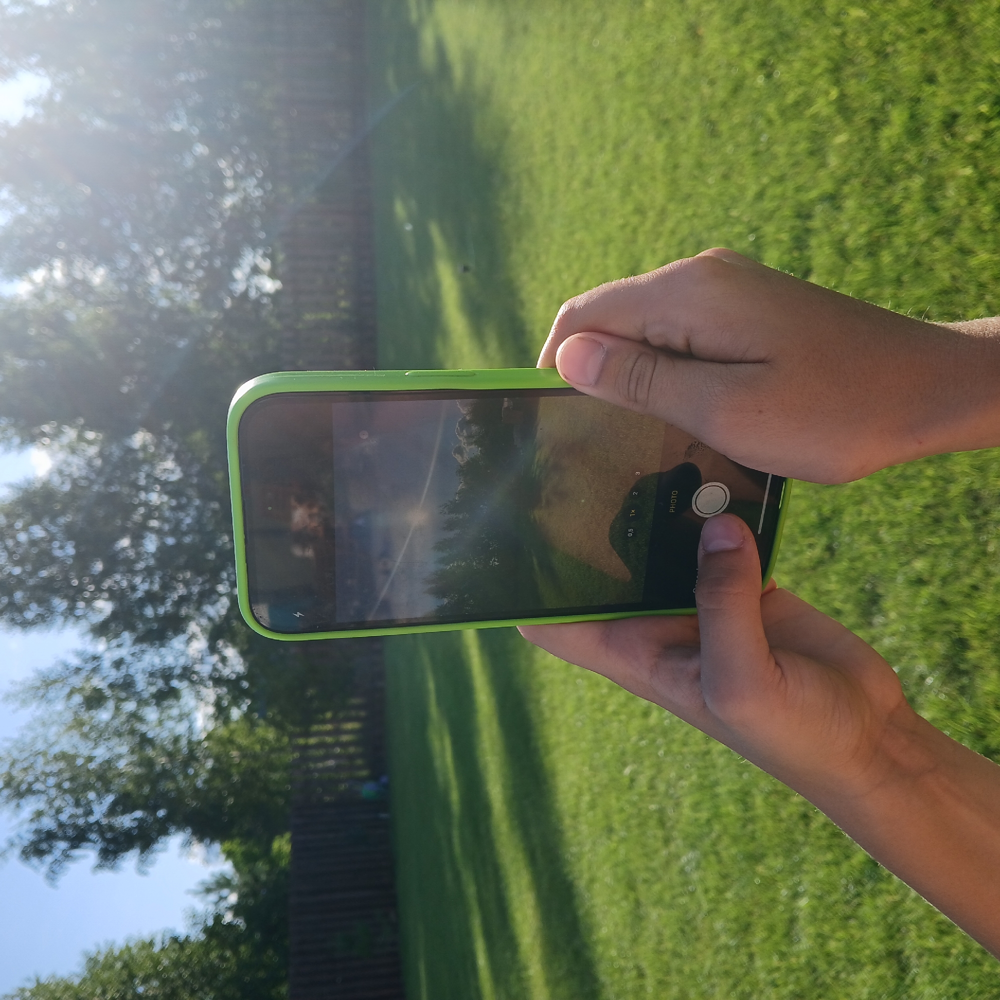 A person is holding a smartphone with a bright green case in their right hand, aiming it towards a sunlit backyard. The phone screen displays a camera interface, with a bright sun and trees visible through the lens, suggesting the photo is being taken outdoors during the day. The background consists of a green lawn, a wooden fence, and more trees, illuminated by the sun, which is casting long shadows. The overall environment appears to be a residential backyard in the city of Samarsky, Russia.
