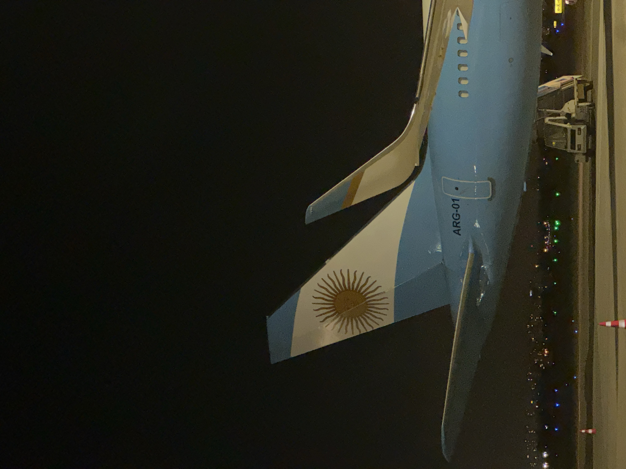 An upward, medium shot depicts the tail fin of the ARG-01 aircraft against a dark, nighttime background. The tail fin is painted with the Argentinian flag, featuring a white field and blue stripes along the top and bottom edges. A golden sun emblem is displayed in the center of the white field. The plane’s body has ARG-01 printed on it, is blue and runs along the right of the frame, extending beyond the top edge. A ramp connects to the plane. Runway edge lights and city lights stretch along the bottom right of the frame, indicating an airport environment at night. The location appears to be at night in Yagel, Israel.