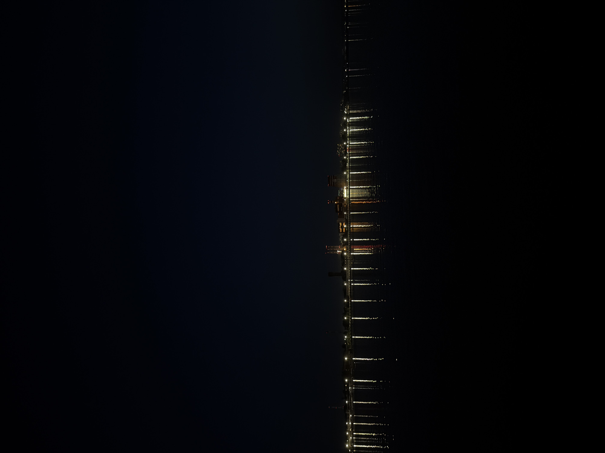 A vertical aerial shot depicts a nighttime cityscape, possibly the city of Izhevsk, Russia. A series of evenly spaced lights extend in a straight line, likely along a body of water like a river or lake. These lights are mirrored by their reflections in the water, indicating a calm surface. The surrounding areas on either side of the illuminated stretch are cloaked in darkness, suggesting a sparsely populated or undeveloped area or possibly an obscured view. The lights suggest an industrial or developed area.