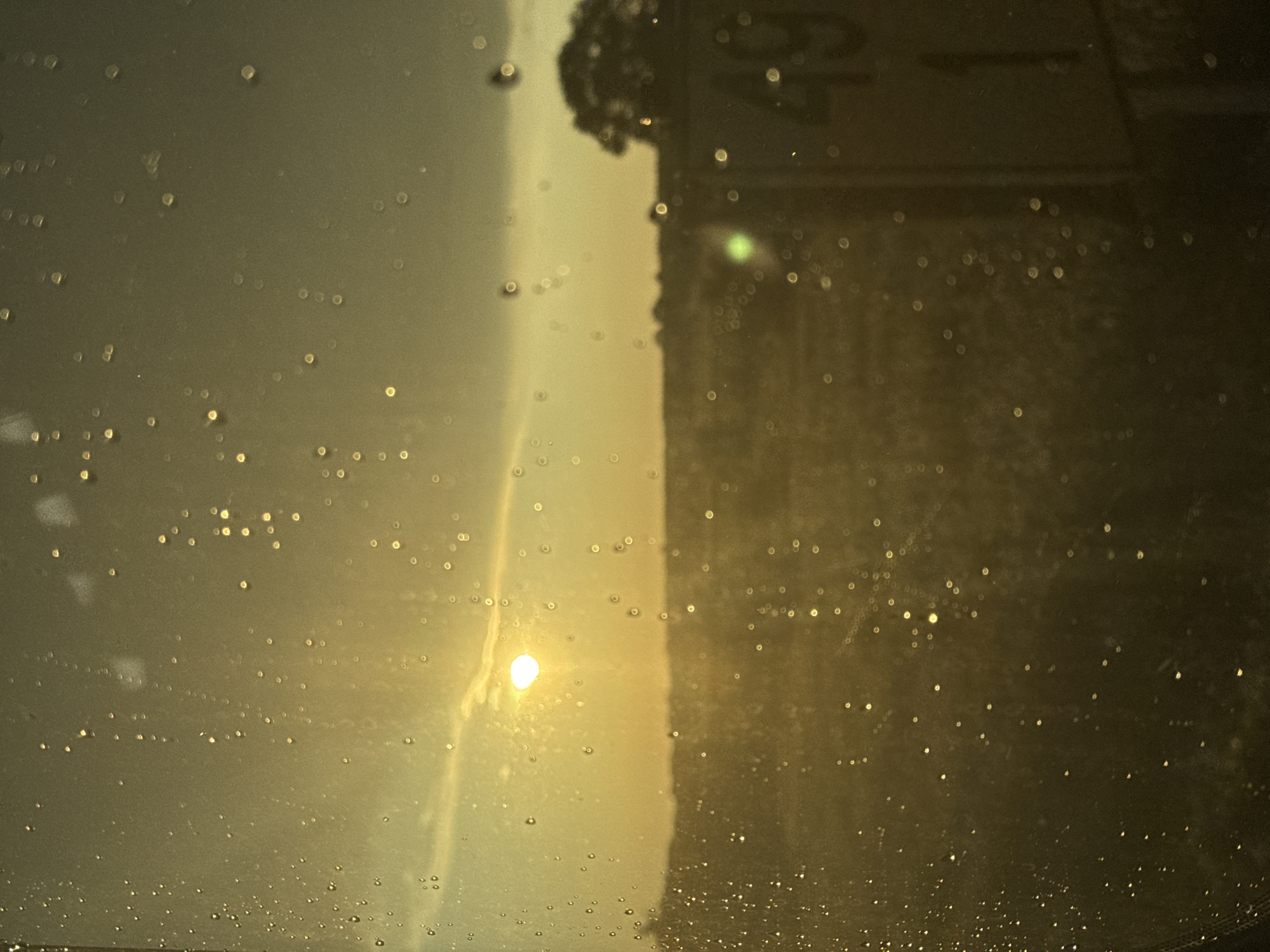 The image is a photograph taken through a wet window or glass surface, showing a landscape scene at sunset. The foreground is dominated by numerous water droplets of varying sizes, which create bokeh effects and obscure the view to some extent. The overall color palette is warm, with golden hues from the setting sun.
In the middle ground, a flat, open field is visible, stretching towards a distant horizon. The field appears to be covered in vegetation, possibly crops or grass, and is cast in a soft, diffused light. There are some indistinct shapes of trees or bushes along the horizon line.
To the right of the frame, a signpost is partially visible. It appears to be a white post with black or dark numerals. The numerals "49" are clearly visible on the upper part of the sign, and a "1" is visible below it. This signpost is likely a mile marker or similar roadside indicator.
In the background, the sun is setting, creating a bright, circular glow in the sky. Wispy clouds are present, catching the golden light. The sky transitions from a bright yellow-orange near the sun to a darker, muted tone towards the top of the frame. The overall lighting suggests late afternoon or early evening.
The image does not contain any people or animals. The scene depicts a rural or semi-rural environment. The context provided suggests this location is in Nowe Tłoki, Poland.