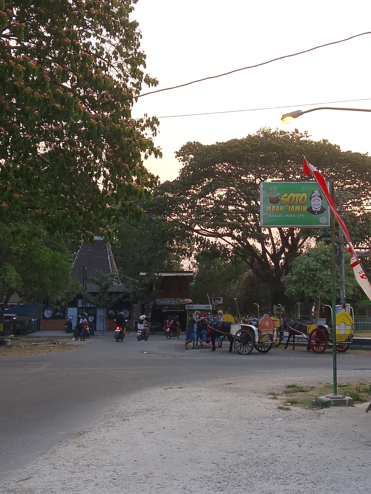 The image depicts a street scene in the city of Balun, Indonesia, likely during the late afternoon or early evening given the warm lighting. In the foreground, a paved road transitions to a rougher, gravelly shoulder. Several horse-drawn carriages, known as delman, are parked or moving slowly. Some of these carriages are elaborately decorated with red and yellow accents, while others are simpler. People are visible near the carriages, some appearing to be drivers or passengers.
Further back, on the left side of the road, is a building with a distinct, tiered roof, suggesting traditional architecture. Scooters and motorcycles are parked and in motion in front of various establishments lining the street. A prominent sign, predominantly green, reads "SOTO MBAH JAMIN" with a cartoonish illustration of an elderly woman. Red and white flags, possibly the Indonesian flag, are displayed on poles. Trees, some with blooming flowers, flank the scene, contributing to a semi-tropical atmosphere. The sky in the background is a pale, hazy orange, indicating a sunset or sunrise. A street light is illuminated, casting a warm glow. The overall impression is one of a bustling, local street with elements of both modern transportation (scooters) and traditional modes of transport (delman).