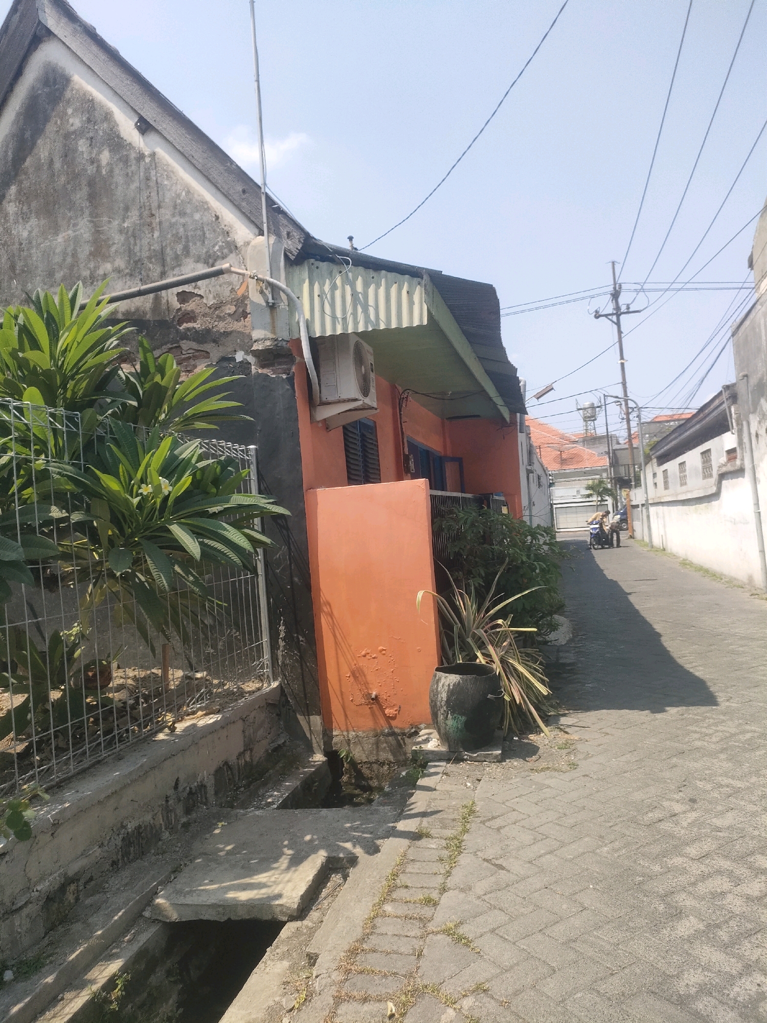 The image depicts a narrow, paved alleyway in Surabaya, Indonesia, lined with residential buildings and vegetation under a clear blue sky. On the left, a dilapidated house with a weathered grey facade and a sloping roof is visible. In front of it, a lush green plant with broad leaves and white flowers grows behind a wire fence. An air conditioning unit is mounted on the orange wall of the adjacent building, which features a green-trimmed overhang. A drainage channel runs along the left side of the alley.
The alley continues into the distance, with more houses and buildings on either side. A motorcycle with two occupants is seen in the mid-ground of the alley, moving away from the viewer. Overhead, a network of power lines crisscrosses the sky, supported by utility poles. The ground is paved with interlocking bricks, and patches of grass grow along the edges. The overall impression is of a residential area in a densely populated urban environment, characterized by a mix of older, well-maintained, and aging structures, with evidence of everyday life unfolding in the street.