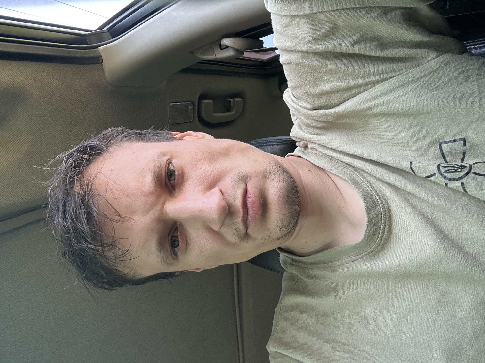 A selfie taken by a man inside a car. The man is looking directly at the camera with a neutral expression. He has short, dark hair that appears messy and a light stubble on his chin. He is wearing a light-colored t-shirt with a graphic design on the left side of his chest. The interior of the car is visible, including the car's headliner, a grab handle, and part of the window frame. The scene suggests the man is in Makiv, Ukraine.