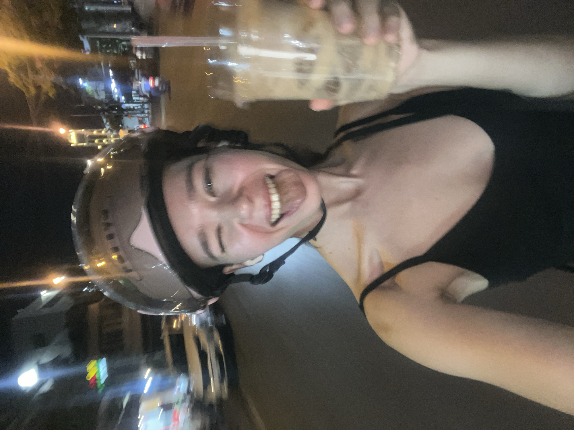 An image, originally taken in portrait orientation and rotated, captures a candid moment at night in Nam Nha Trang Ward, Vietnam. The primary subject is a young person, likely female, seen from the chest up, smiling broadly and looking directly at the camera. Her dark hair is visible, and she wears a dark-colored top with thin straps.
A light-colored helmet with a clear visor and chin strap is on her head, suggesting she is on a scooter or motorcycle. In her right hand, she holds a clear plastic cup containing a light-colored, possibly iced, beverage with a straw. Her wide, joyful smile, revealing her teeth, conveys a sense of happiness and excitement.
The background is dark and out of focus, indicating nighttime and motion blur. Numerous artificial lights from streetlights, buildings, and possibly other vehicles create streaks and glowing spots, characteristic of an urban or semi-urban environment at night. The overall impression is one of enjoying a casual outing or ride in the evening. No discernible text is visible on any signs or objects in the image.