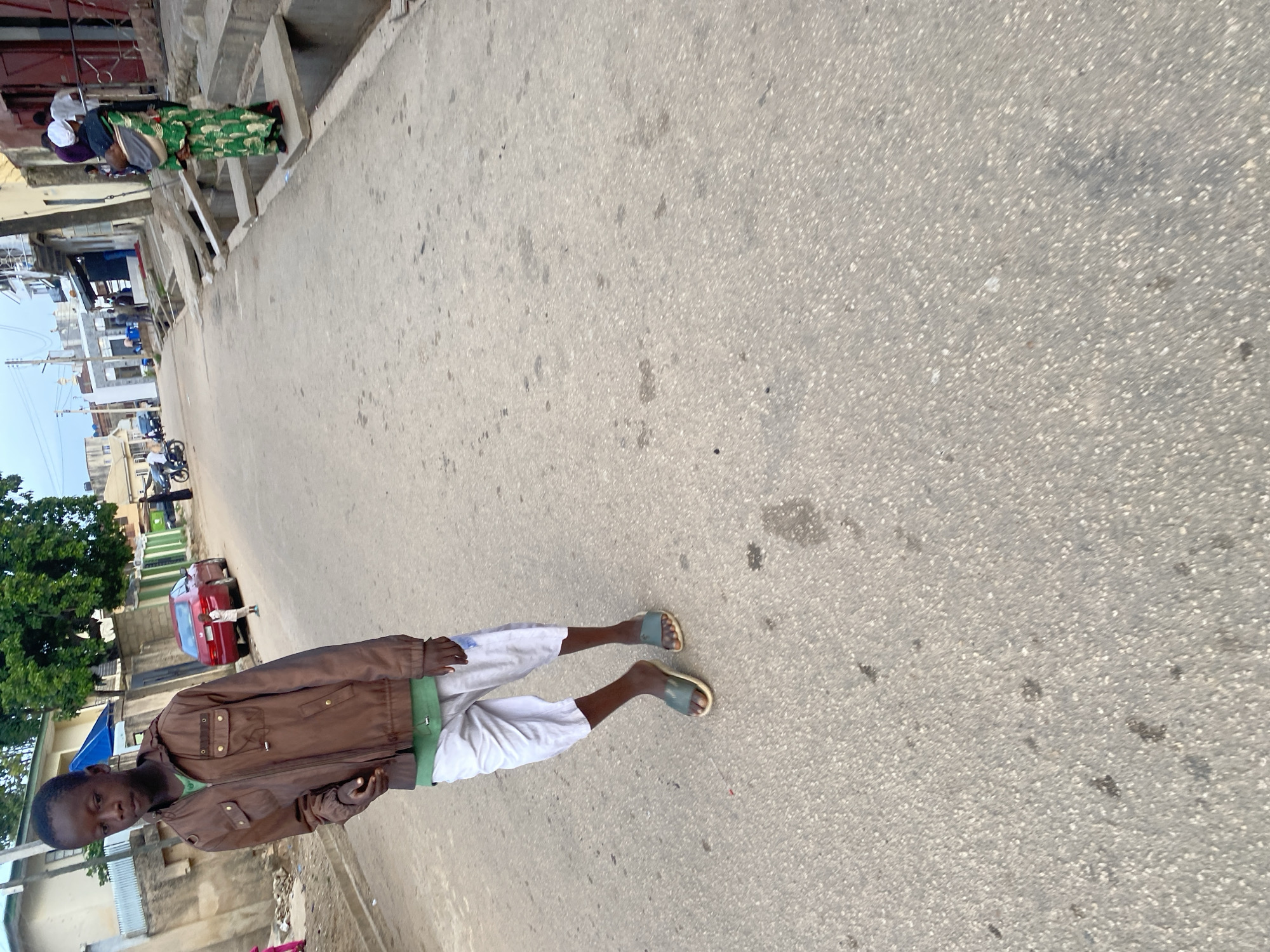 A young boy, likely a teenager, stands on an asphalt road in Jos, Nigeria. He is wearing a brown jacket over a green t-shirt and loose-fitting white shorts. His feet are clad in dark-colored slides. The boy is positioned in the foreground, facing slightly towards the viewer and to the left. Behind him, the road stretches into the distance, flanked by buildings and a drainage system. A red sedan is parked on the right side of the road, and another child can be seen near it. Further down the road, several people are visible, including a woman carrying a baby on her back, dressed in a vibrant green patterned dress. Motorbikes and other vehicles are also present in the background, indicating a typical urban street scene. The sky is overcast, suggesting it might be late afternoon or early morning. The overall impression is a candid snapshot of everyday life in a Nigerian city.
