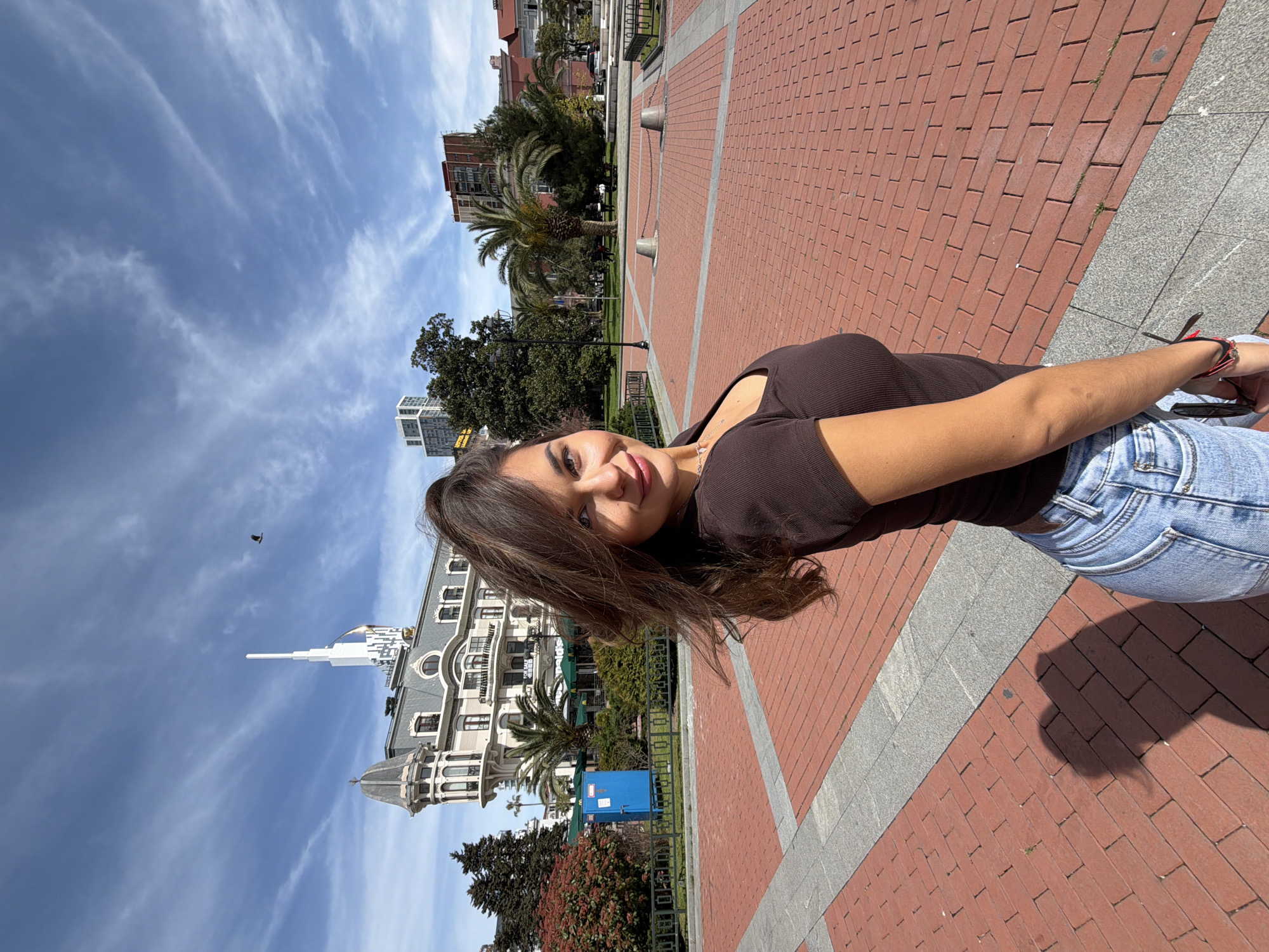 The image shows a young woman in a brown ribbed top and light blue jeans standing on a brick walkway in Batumi, Georgia. Behind her is a view of the city featuring distinctive architecture, including a building with a white spire and a domed roof. Palm trees and other greenery are visible, adding to the urban landscape. The sky is blue with scattered clouds. The photograph appears to be taken during the daytime under sunny conditions.