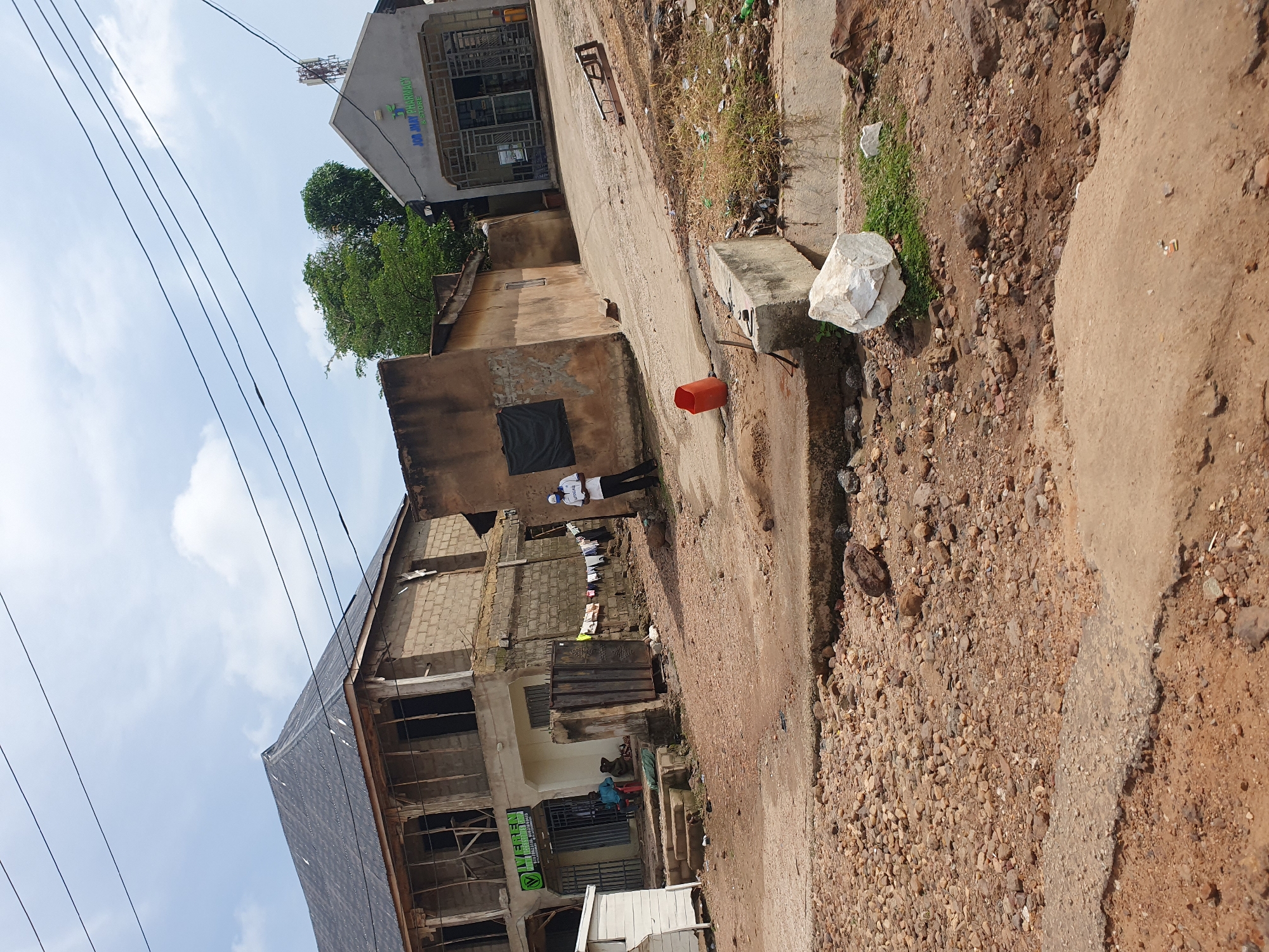 The image shows a street scene in Lokoja, Nigeria. A partially constructed building with a dark roof stands on the left. In front of it is a store with a green sign that reads "VEREN MOBILE PHONE SHOP". Two men are seated on a step in front of the store, one in blue and the other in brown. A metal gate and some hanging clothes are visible to the right of the store. Further to the right, a grey building with a sign that reads "JOB JUMAY PHARMACY & STORE" is visible, with a cell tower on its roof. A man in a white shirt and blue cap stands near the pharmacy. The ground is a mixture of dirt and gravel, with some concrete structures and debris scattered around. The sky is partly cloudy.