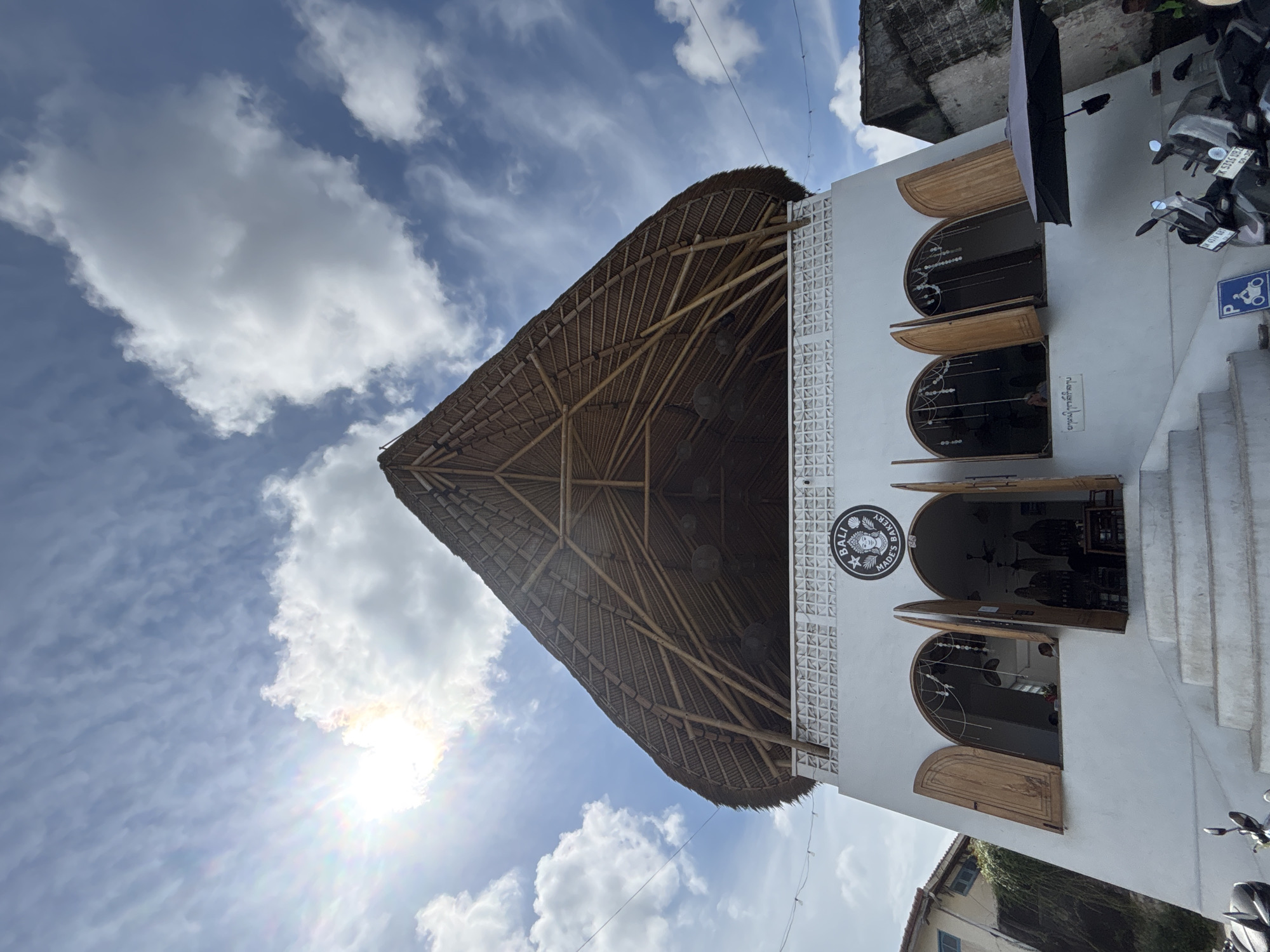 This image depicts the exterior of a building in Kuta Selatan, Indonesia, likely a restaurant or shop, with a distinctive thatched roof supported by visible bamboo beams. The building is white with arched windows and wooden shutters. A sign above one of the arched entrances reads "BALI MADE'S BAKERY". The sky is blue with scattered white clouds and the sun is partially visible, creating lens flare in the upper left portion of the image. In the foreground, two motorcycles are parked, and a blue parking sign is visible. The overall scene suggests a bright, sunny day in a tropical location.