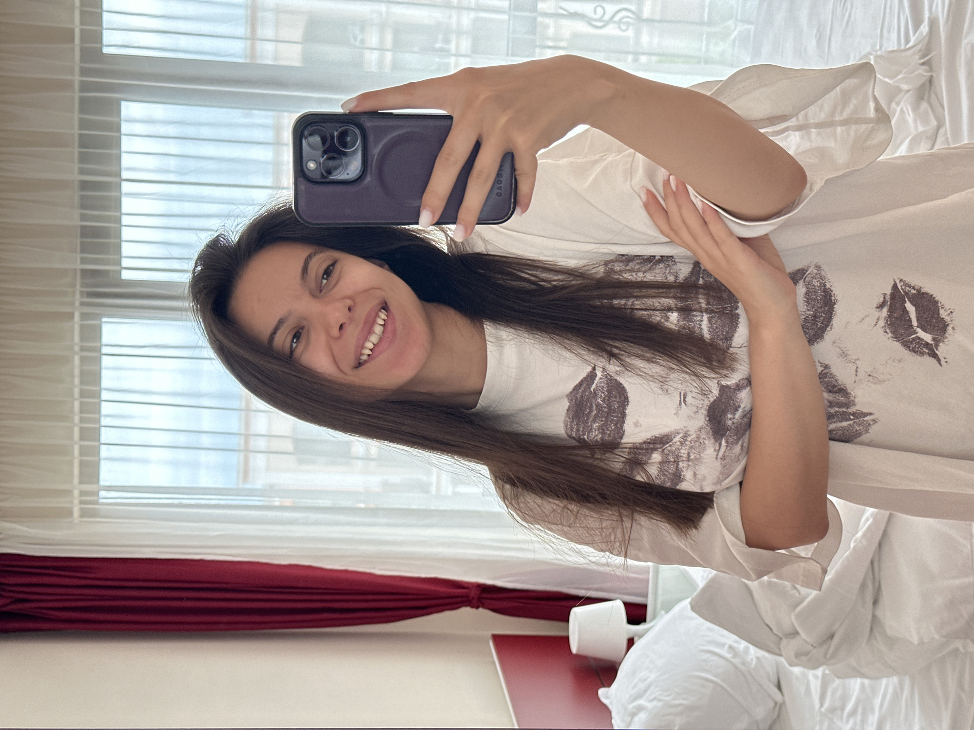 A young woman with long dark hair takes a selfie in a bedroom. She is wearing a white t-shirt with a dark graphic print and is holding a dark-colored smartphone in her right hand, angled to capture her reflection in a mirror. Her left arm is crossed over her chest, and her right arm is bent at the elbow. She is smiling with her teeth showing.
The background reveals a neatly made bed with white bedding and pillows. To the left, there is a red headboard and a small white lamp on a bedside table. Heavy red curtains are drawn to the left of a large window covered by sheer white curtains. The window appears to overlook a residential area with balconies visible in the distance, suggesting an urban or resort setting. The overall impression is of a bright, daytime scene in a contemporary hotel room or apartment.
This scene is located in Sveti Vlas, Bulgaria.