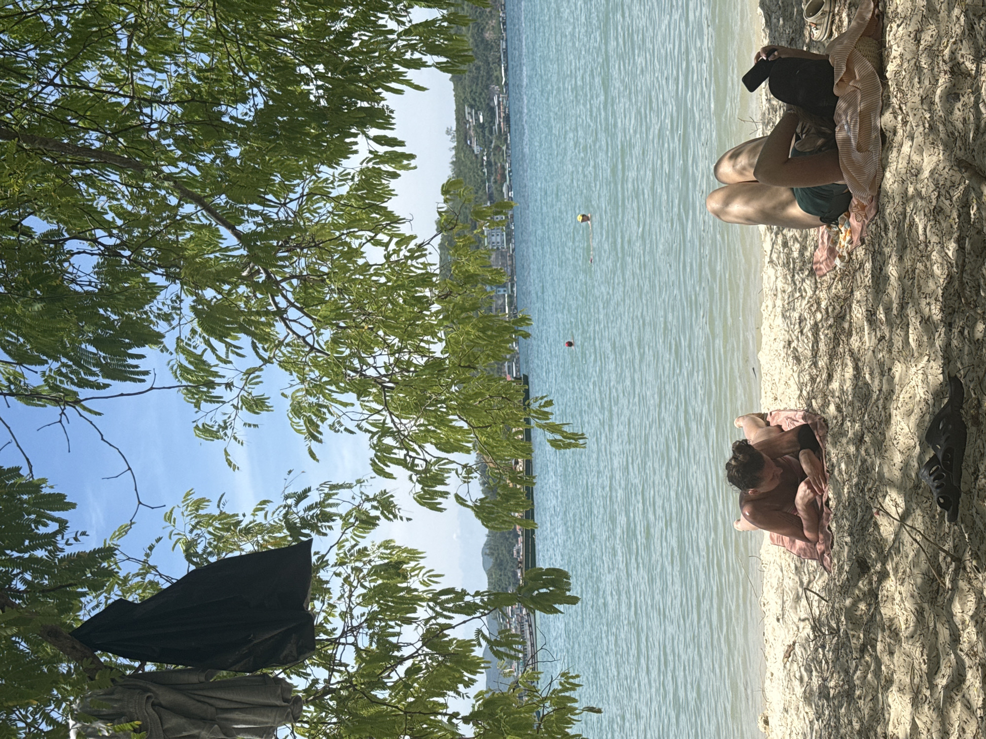 Under the shade of a tree with lush green foliage, two individuals are relaxing on a sandy beach by the turquoise waters of Nha Trang, Vietnam. The person on the left, facing down, is lying on a towel, engrossed in a book. The person on the right is lying on their back, holding a phone and wearing a hat. The ocean stretches out to the horizon, with a few distant boats visible. Buildings and hills line the coastline in the background. A black bag and a grey garment hang from a tree branch in the foreground, adding to the casual beach scene.