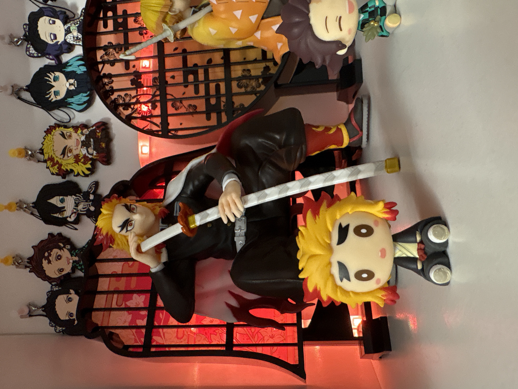 The image shows a display of Demon Slayer anime merchandise. Two main figures depict the character Rengoku. One figure is posed dynamically, holding a sword. Another figure is a smaller, stylized chibi version. The background features a lattice design with red LED lighting. Hanging on the side are keychains of various Demon Slayer characters. The setting is not identifiable as being in Plovdiv, Bulgaria, as there are no visual clues or context to suggest that location.