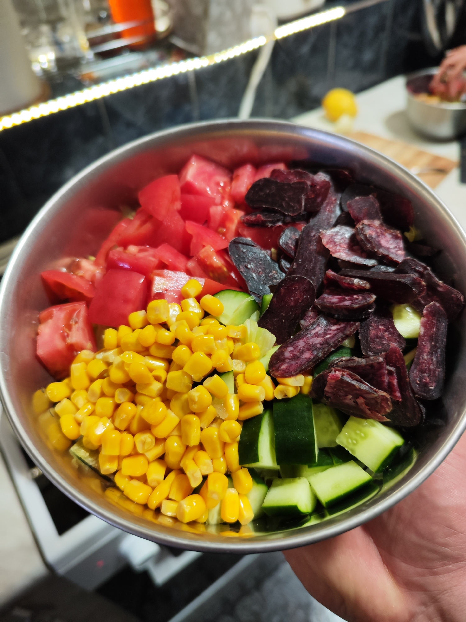 The image displays a close-up, high-angle view of a stainless steel bowl filled with various salad ingredients, held by a person's hand in the lower right. The bowl's contents are distinctly partitioned: bright red diced tomatoes occupy the top left, vibrant yellow corn kernels are positioned in the bottom left, dark green diced cucumbers are in the bottom right, and slices of dark reddish-brown dry sausage, resembling sujuk or another cured meat, fill the top right section.
The background reveals an indoor kitchen setting. A dark tiled wall stretches across the upper part of the frame, illuminated by a horizontal strip of warm-toned LED lighting. Below this, a light-colored countertop features a yellow lemon, a wooden cutting board, and a small metal bowl. A second person's hand is partially visible near the small bowl, suggesting collaborative or ongoing food preparation. Partially visible beneath the main bowl is a section of a white and black kitchen appliance, possibly a stovetop or oven control panel.
The scene captures a moment during food preparation, likely the assembly of a salad. The artificial lighting from the LED strip suggests the photograph was taken indoors, possibly in the evening. No discernable text is present within the image. The location context for this activity is Burgas, Bulgaria.