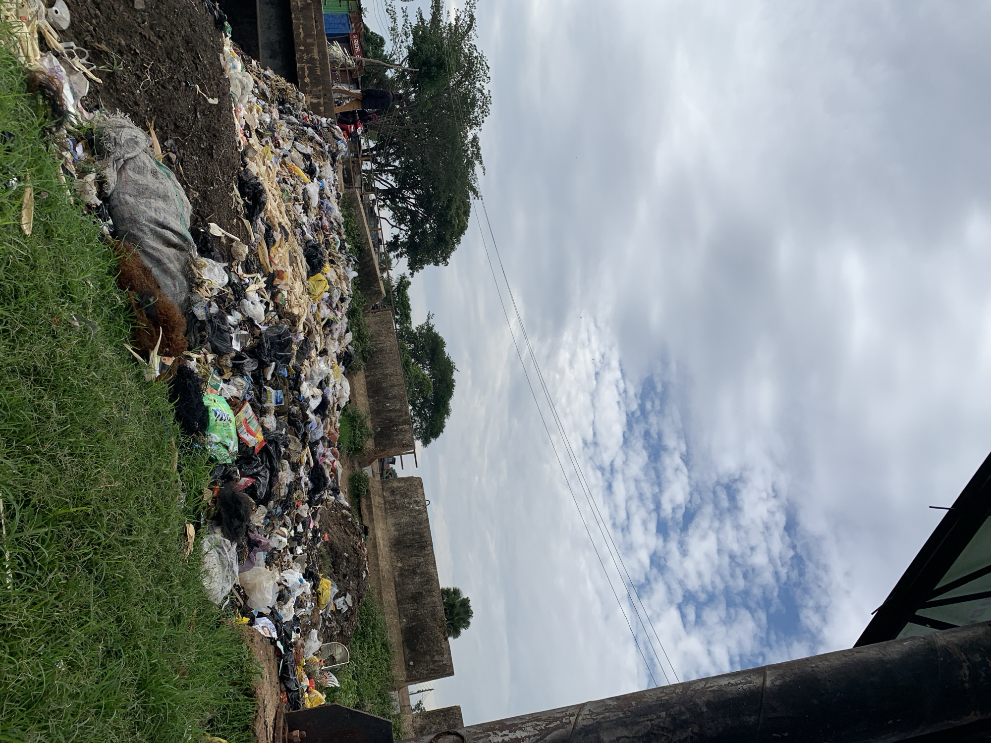 This image, taken in Rafin Par, Nigeria, depicts an extensive accumulation of uncontained refuse forming a large mound alongside a road or embankment. The sky above is largely overcast with grey and white clouds, revealing patches of blue sky, particularly towards the upper right.
In the foreground, lush green grass covers the lower right portion. A thick, dark, cylindrical column, possibly a utility pole or structural support, occupies the far right side of the frame, extending upwards and partially out of view. Attached to this column, in the upper right corner, is a dark, angular structure with a green underside, suggesting an overhead canopy or architectural element.
The central and lower-left midground is dominated by a massive pile of diverse garbage. This refuse consists predominantly of discarded plastic bags in various colors, including numerous black, white, and yellow bags, along with fabric scraps, soiled materials, and other unidentified debris. The garbage rests upon dark soil, which is exposed in several areas, particularly on a slope on the lower left.
In the background, a raised concrete structure or road is visible above the refuse pile. On this elevated platform, towards the left, two individuals are present. One person, dressed in dark attire and light-colored trousers, stands near a red motorcycle, facing towards the right. The second individual is partially obscured behind the first. Further left in the background, a small structure with a blue base and a green upper section displays partial text, "SERCOFEL S". Multiple large trees with dense green foliage are prominent in the left and central background. Utility lines are stretched horizontally across the mid-sky. Towards the right background, several substantial grey concrete barriers, resembling road dividers, are aligned horizontally. A single palm tree is discernible behind one of these barriers.