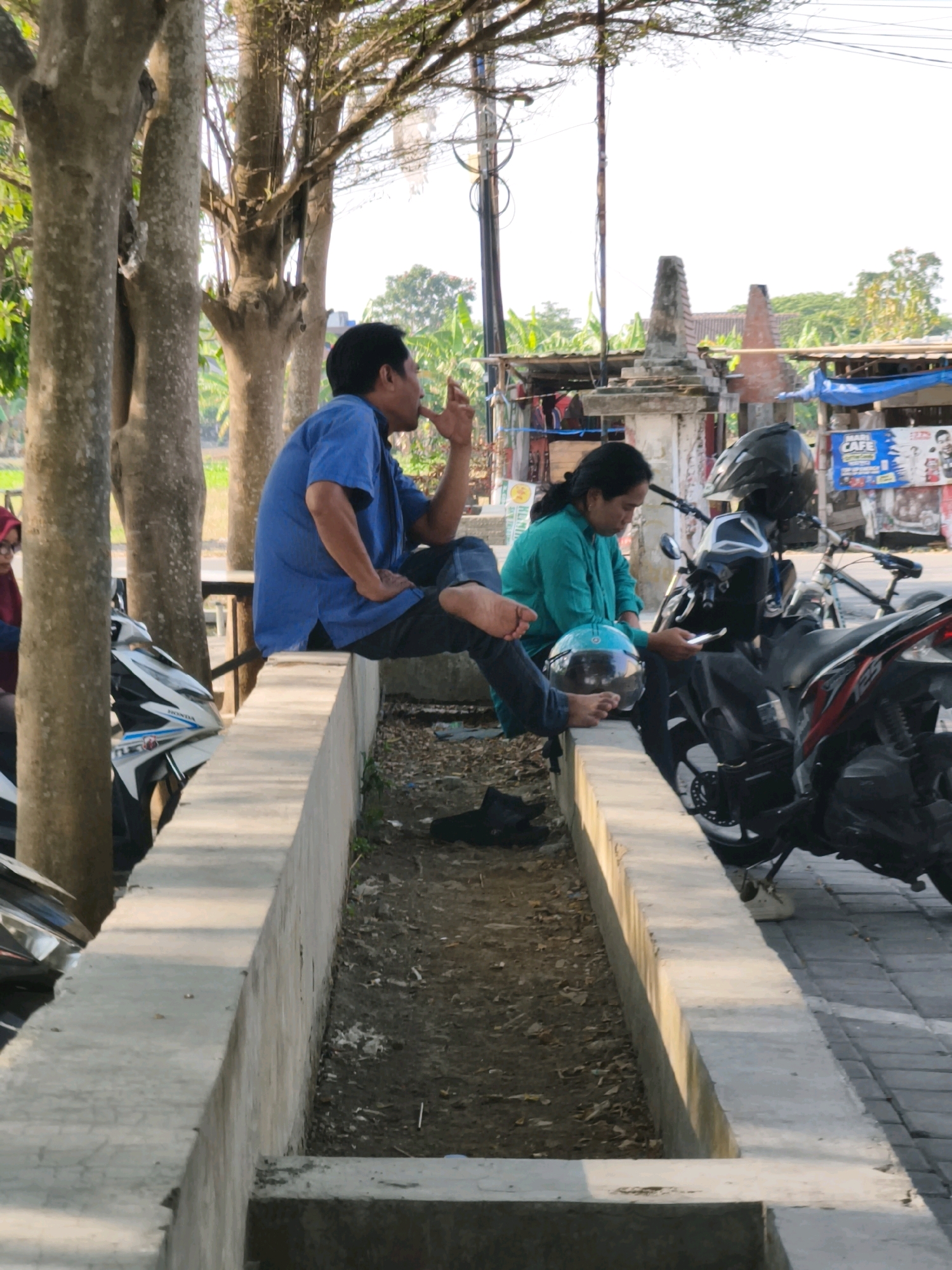 Two individuals are present in an outdoor urban setting, likely near a road or pathway in Desa Dibal, Indonesia. On the left, a man wearing a blue short-sleeved shirt and dark pants is seated on a concrete ledge, with his left leg bent and right leg extended. He appears to be smoking and is looking to his right. To his right, a woman in a teal long-sleeved shirt and dark pants sits on the same ledge, holding a light blue helmet in her left hand and a white smartphone in her right hand. She is looking down at the phone. Several motorcycles are parked behind them, with one black helmet resting on a motorcycle. The environment includes trees with visible branches and leaves, a pole with wires in the background, and a building with a corrugated roof. A concrete structure, possibly a drainage ditch or planter, runs along the foreground. The ground is covered with dirt, leaves, and debris. The lighting suggests it is daytime, with shadows cast on the concrete ledge.