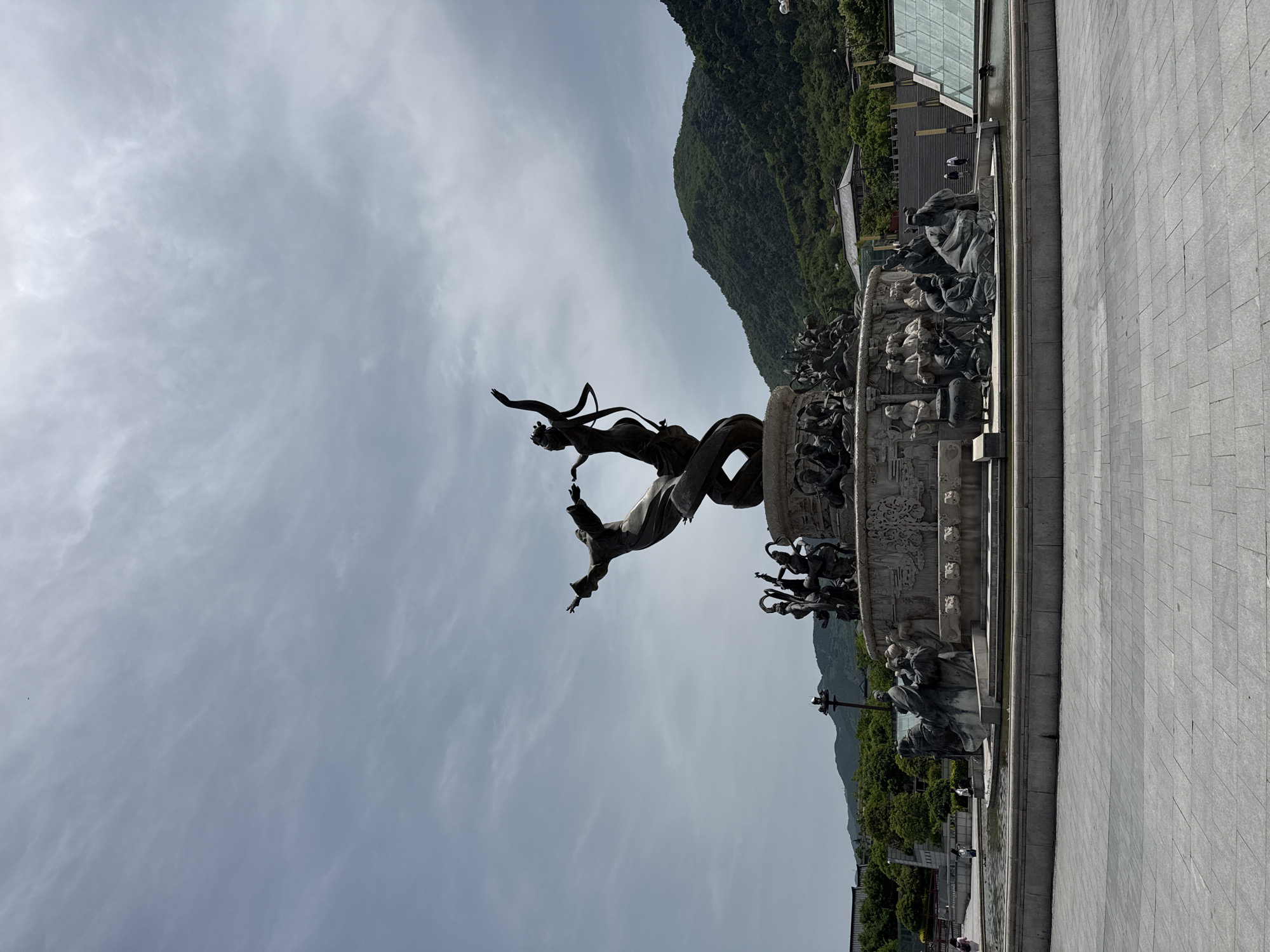 The image depicts a large, bronze sculpture in Lintong District, China. The sculpture appears to be a fountain, featuring a central figure leaping or flying through the air, intertwined with what seems to be a dragon or serpentine form. The base of the sculpture is adorned with intricate carvings and multiple smaller figures.
The background shows a lush, green mountain rising sharply behind the sculpture, and a modern, glass-walled building is visible on the right side of the frame. The foreground consists of a paved plaza or square with a light gray color, suggesting the sculpture is located in a public space. The sky is overcast with light gray clouds.