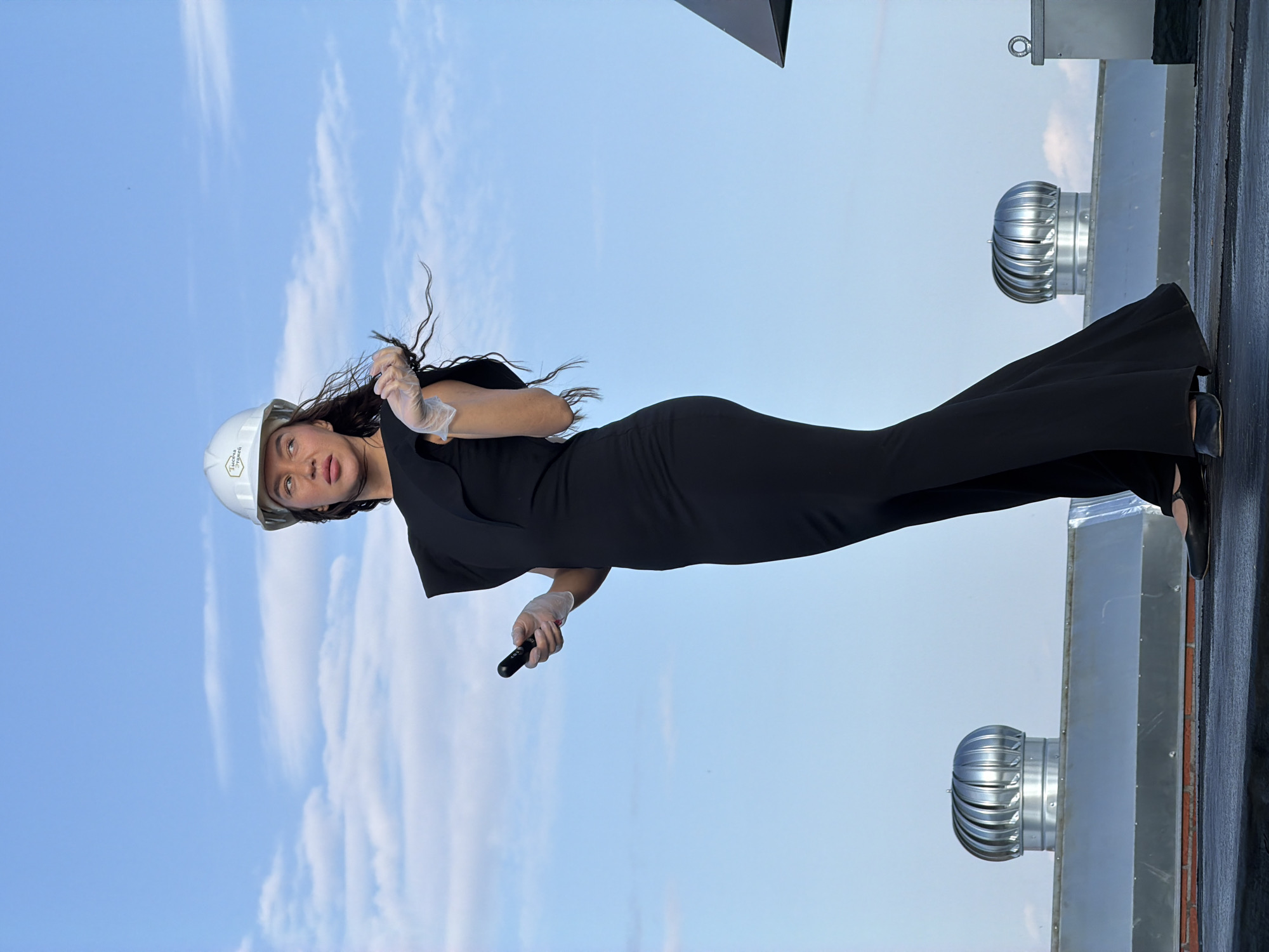 A woman wearing a black dress and a white hard hat stands on a rooftop in Chaikovsky, Russia. She is also wearing clear gloves and holding a black remote in her right hand. The woman has long, dark, wavy hair. The background is a bright blue sky with some wispy clouds. On the rooftop, there are several metal vents and a black surface. The woman is looking to her left.