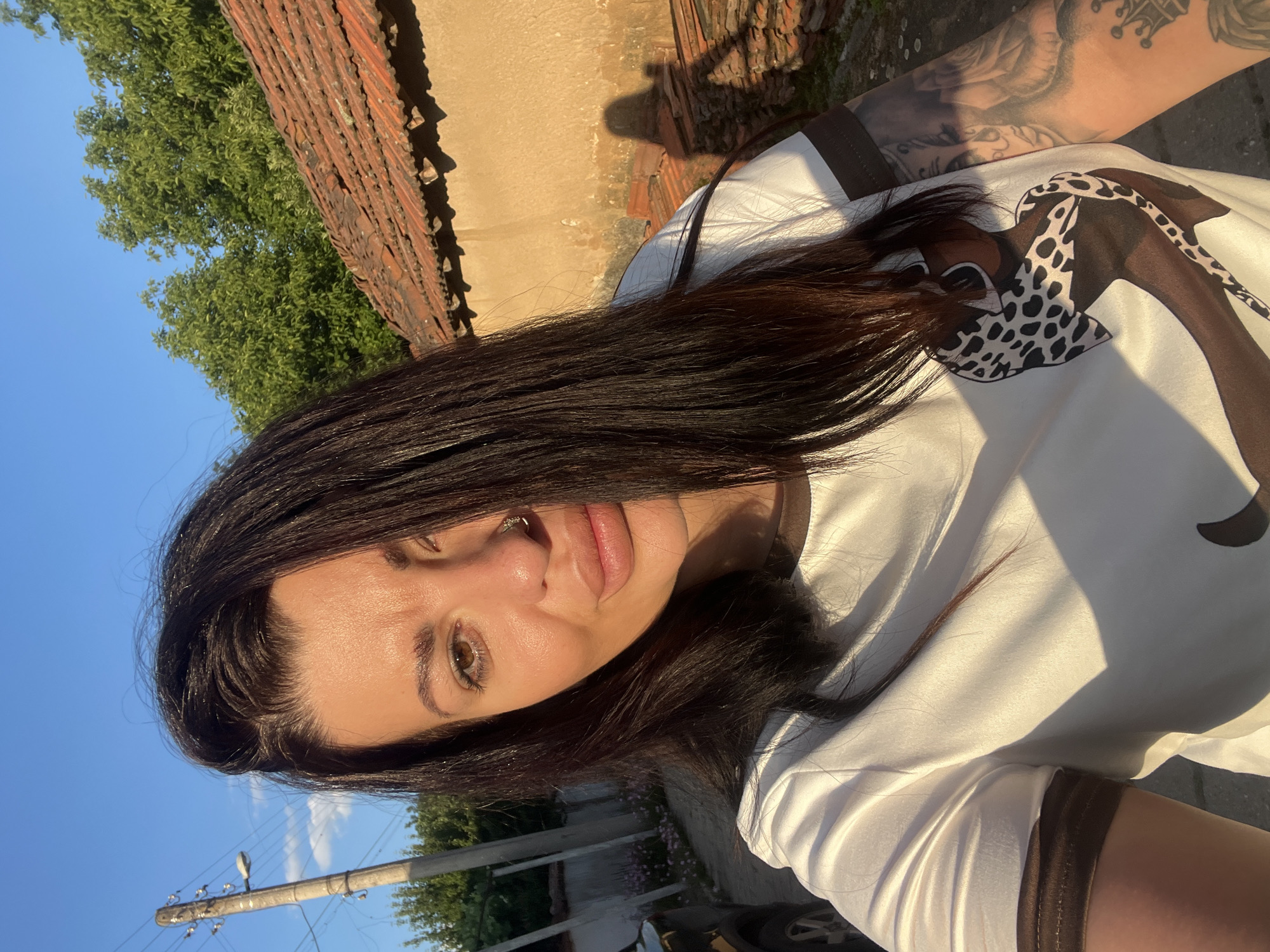 A medium-shot, waist-up selfie of a woman with long, dark brown hair, fair skin, and brown eyes is taken outdoors during golden hour. She is wearing a white t-shirt with dark brown trim on the sleeves and neckline, featuring a graphic print of a stylized figure. A nose ring is visible. Her left arm, which is tattooed, is partially visible in the frame. The background suggests a rural setting in Zelenikovo, Bulgaria, with a shadow of the photographer cast on an ochre-colored wall with a red-tiled roof. A utility pole and trees are also visible. The sunlight is strong, creating a warm glow and distinct shadows.