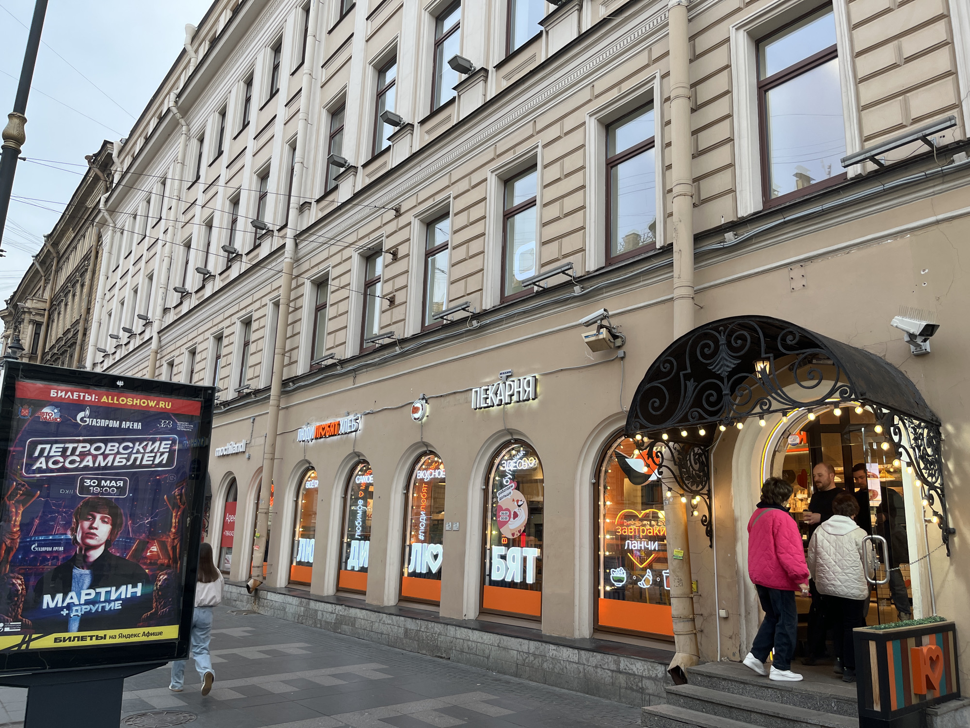The image captures a street scene, likely in Russia given the Russian text, showcasing a bakery storefront and an adjacent advertising billboard. The bakery, named "People Love Bread" (translated), features arched windows displaying images of food items and neon text. Two people are seen entering the bakery, while another person is walking away. The building is a multi-story structure with classic architectural details. The advertising billboard on the left promotes an event called "Petrovsky Assemblies" scheduled for May 30th, featuring an artist named "Martin." The environment is urban, with a gray sky suggesting it may be an overcast day. The sidewalk is paved, and a light pole is visible on the left.