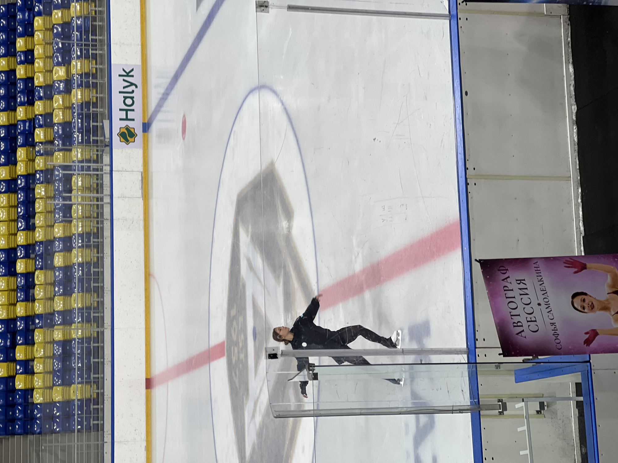 A full-body, slightly angled shot shows a young female figure skater in mid-performance on an ice rink. She is wearing a black athletic suit and white skates, with her arms extended and her body slightly bent as she glides across the ice. The rink features red and blue markings, a large stylized logo in the center, and a "Halyk" advertisement banner visible in the background. Above the rink, tiered blue and yellow seating is visible, suggesting a sporting venue. In the foreground, a poster is displayed, featuring a photo of a woman with her arms raised and text in Russian that reads "AUTOGRAPH SESSION SOFIA SAMODELKINA". The scene is indoors, likely at an ice skating arena. The image is dated as being taken in Almaty, Kazakhstan.