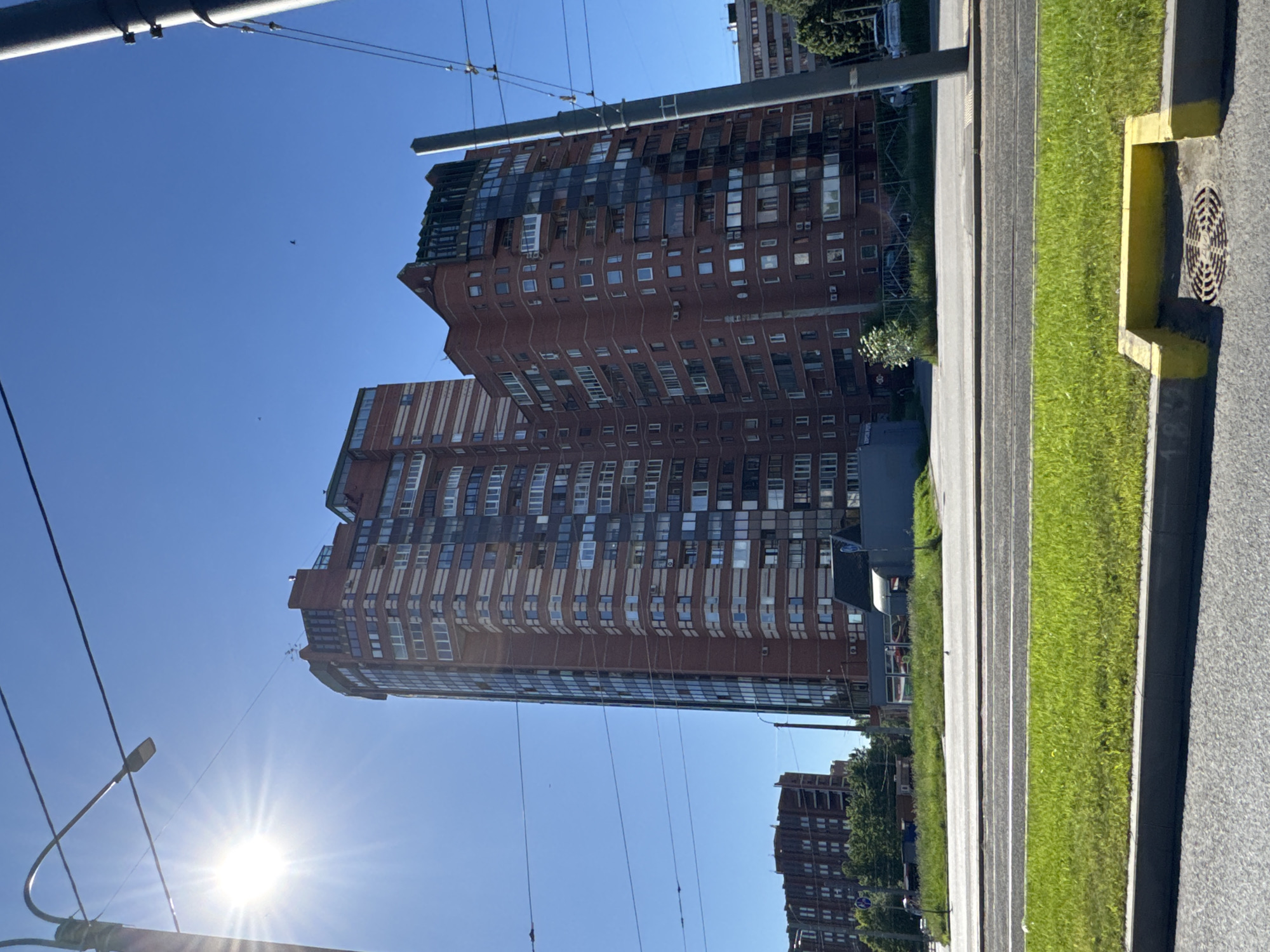 The image depicts an urban street scene in Yukki, Russia, under a clear, bright blue sky with direct sunlight causing a prominent sun flare in the upper left. The primary focus is on two substantial, multi-story residential buildings constructed with red brick. The taller building on the left is curvilinear in design, featuring extensive dark glass facades interspersed with bands of red brick and numerous balconies. The adjacent building to its right is also red brick with dark glass elements, presenting a more angular roofline and various window styles.
In the foreground, a multi-lane asphalt road is visible, featuring a central grassy median strip containing tram tracks. A storm drain with a circular metal grate is present in the bottom right corner of the foreground, bordered by a curb painted yellow. Utility poles with overhead wires traverse the scene, with one prominent dark grey pole on the far right and another with a streetlamp in the upper left. Additional red-brick residential buildings are partially visible further down the road on the left, partially obscured by green trees. Small birds are discernible against the clear sky. No identifiable individuals are clearly visible.