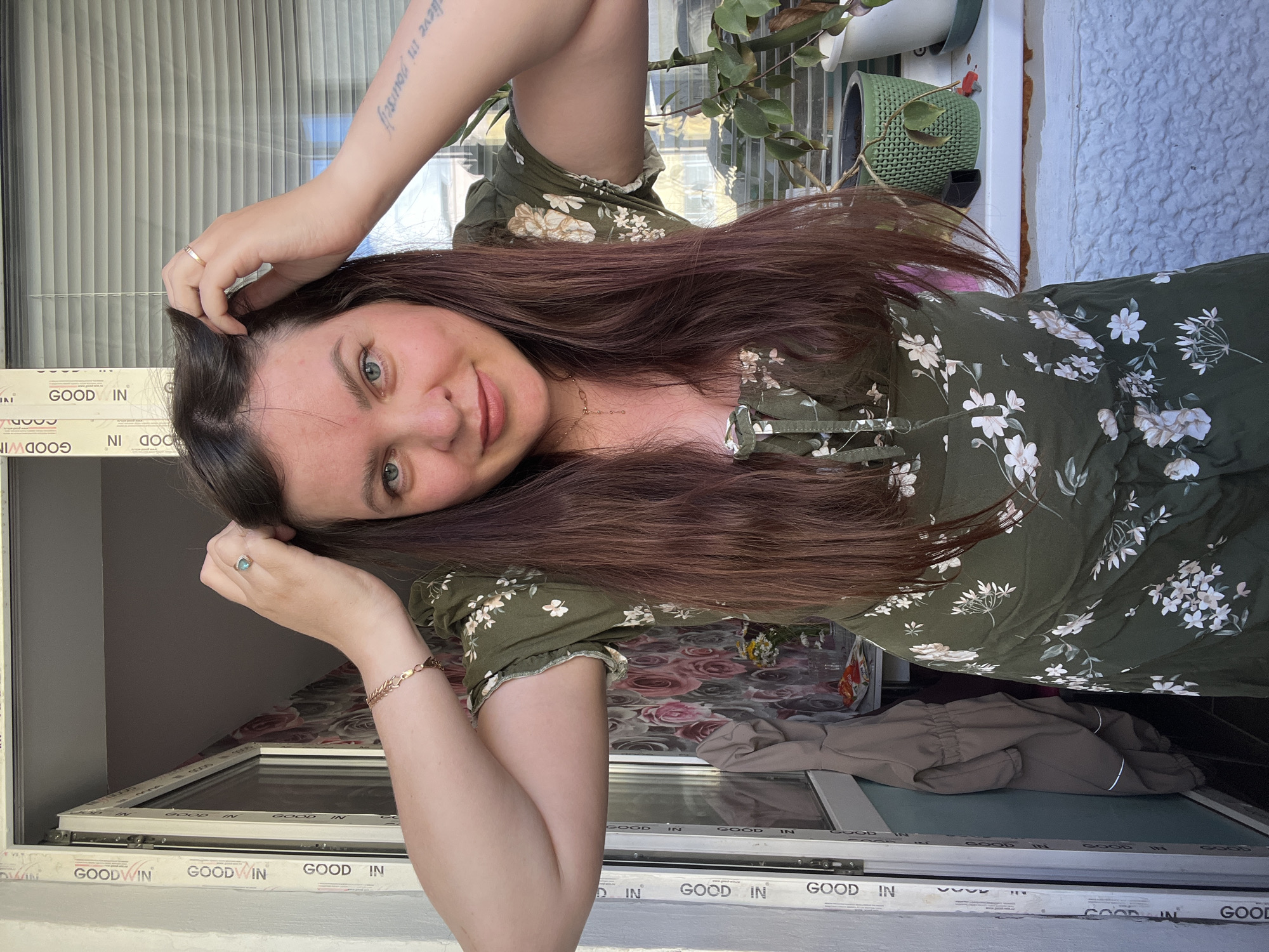 A young woman with long, dark brown hair and blue eyes is standing on a balcony, possibly in the city of Noviy, Russia, as indicated by the "GOOD WIN" branding on the window frame. She is wearing a green dress with a floral print and has her hands on her head, touching her hair. A ring with a blue stone is visible on her left hand. She also has a bracelet on her left wrist. A tattoo is visible on her right forearm. The background shows a window with blinds, a plant in a green pot, and what appears to be a coat hanging on a hook to the left. The overall setting suggests a casual, domestic environment.