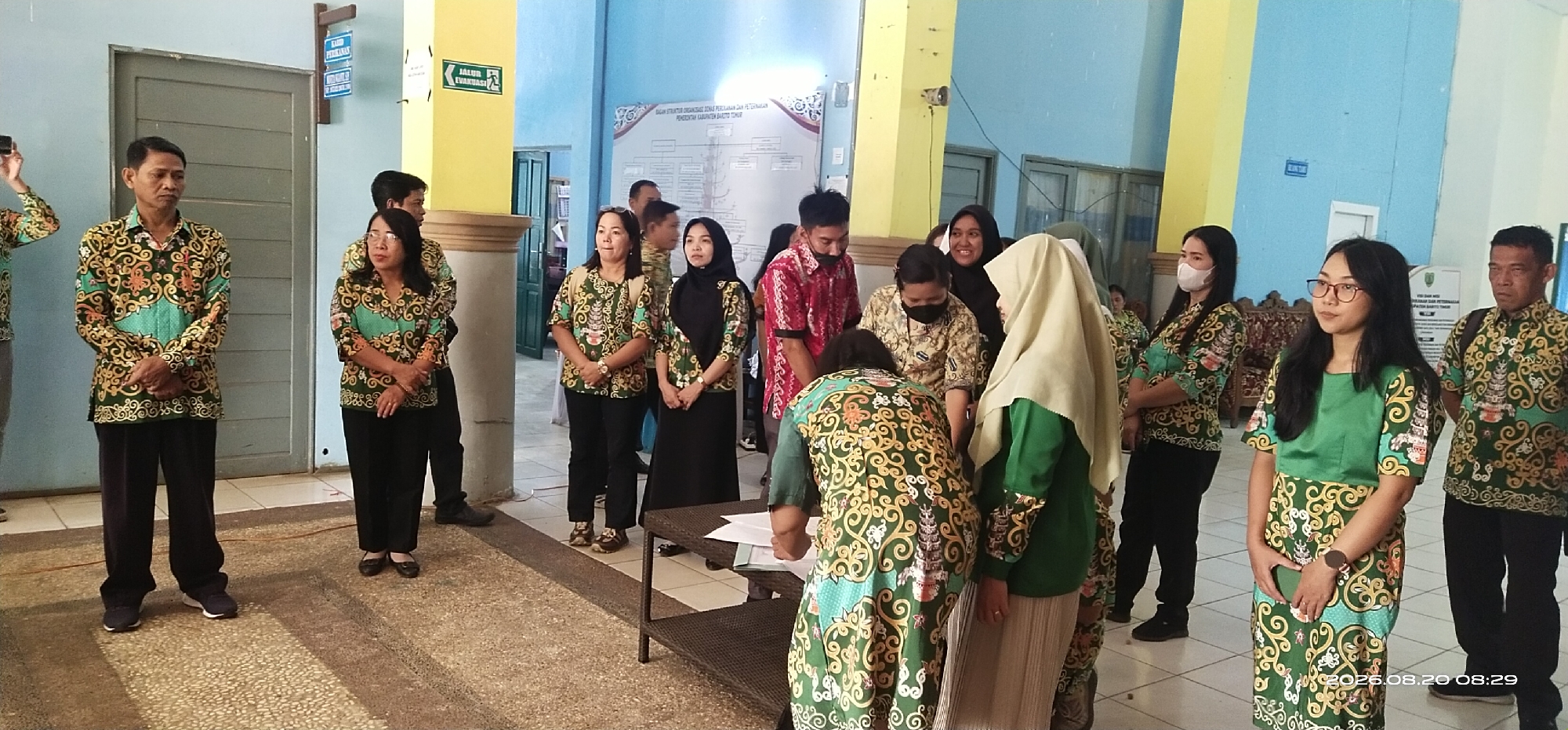 The image depicts a group of people, primarily women, gathered indoors, possibly in a government office or community center in Tamiang Layang, Indonesia. The lighting suggests an indoor environment with a bright, somewhat sterile atmosphere, possibly illuminated by overhead fluorescent lights. The background features light blue walls and yellow pillars, with doors and a bulletin board displaying a flowchart.
The individuals are mostly dressed in matching batik-style shirts, predominantly in shades of green with gold and black patterns. Some are wearing black pants or skirts, while others are in full batik ensembles. One woman in the foreground on the right wears a green top and a matching green skirt with a intricate gold pattern. She has glasses and long dark hair. To her left, another woman in a similar green batik shirt wears a white face mask.
Several people are engaged in conversation or looking towards a table in the center where papers are laid out. A dark-haired woman in a black hijab stands near the table, interacting with others. A man in a red batik shirt is visible on the right side of the frame, looking downwards. Another man in a green batik shirt stands to the far right, his gaze directed forward.
The floor appears to be tiled or concrete with a patterned texture. A dark-colored, multi-tiered shelf unit with papers on it is positioned in the foreground. The overall scene suggests a formal gathering, perhaps a meeting or an orientation. The timestamp at the bottom right corner reads "2026.08.20 08:29".